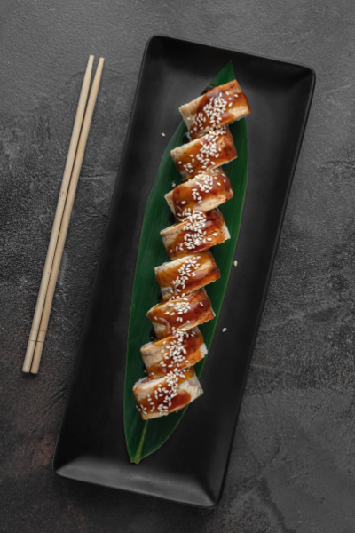 Sushi rolls drizzled with sauce and sprinkled with sesame seeds, served on a green leaf on a black rectangular plate with chopsticks placed alongside.