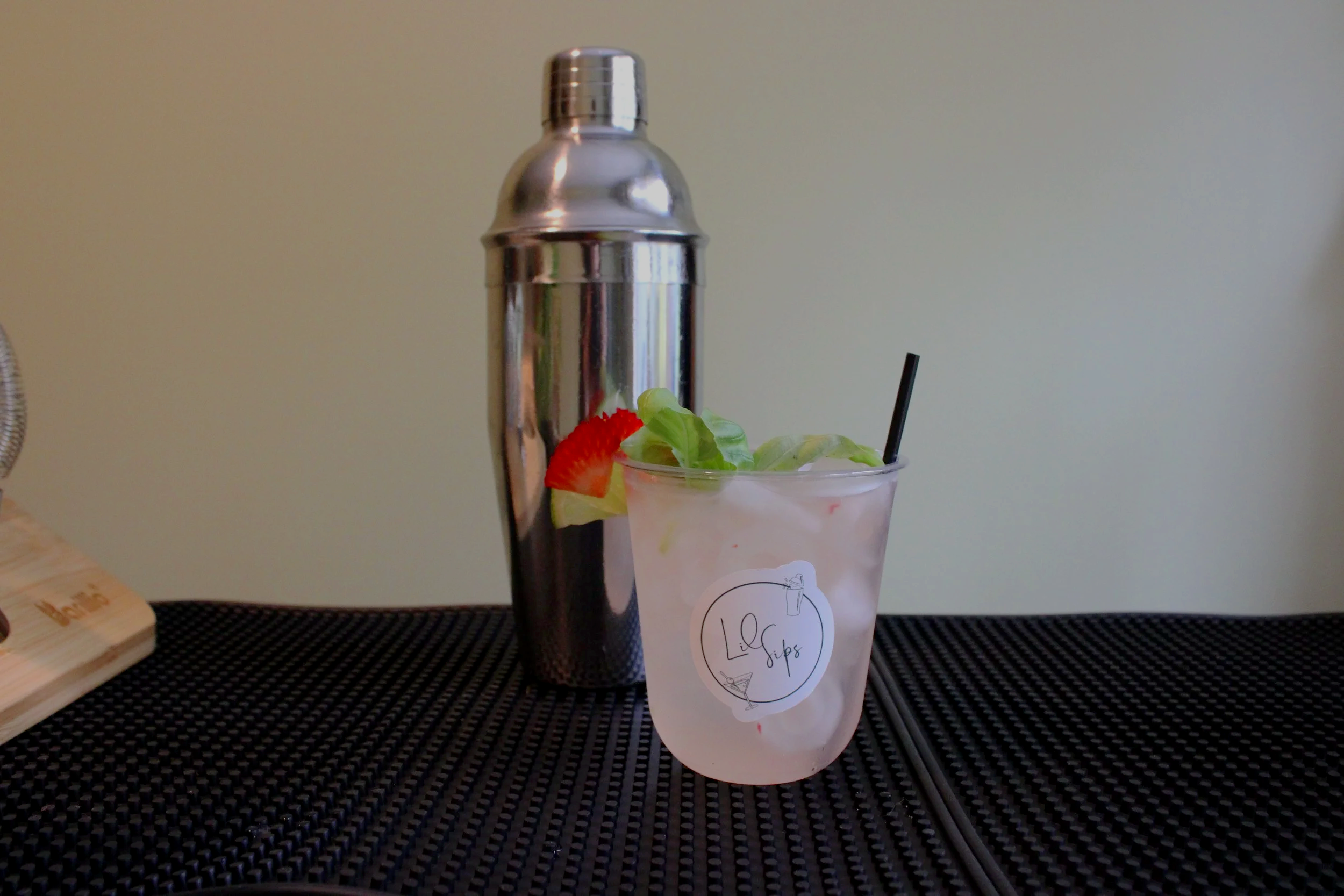 A cocktail glass filled with ice and topped with a lime wedge, a strawberry slice, and a leaf, with a black straw inside. Behind it is a stainless steel cocktail shaker on a black textured surface.