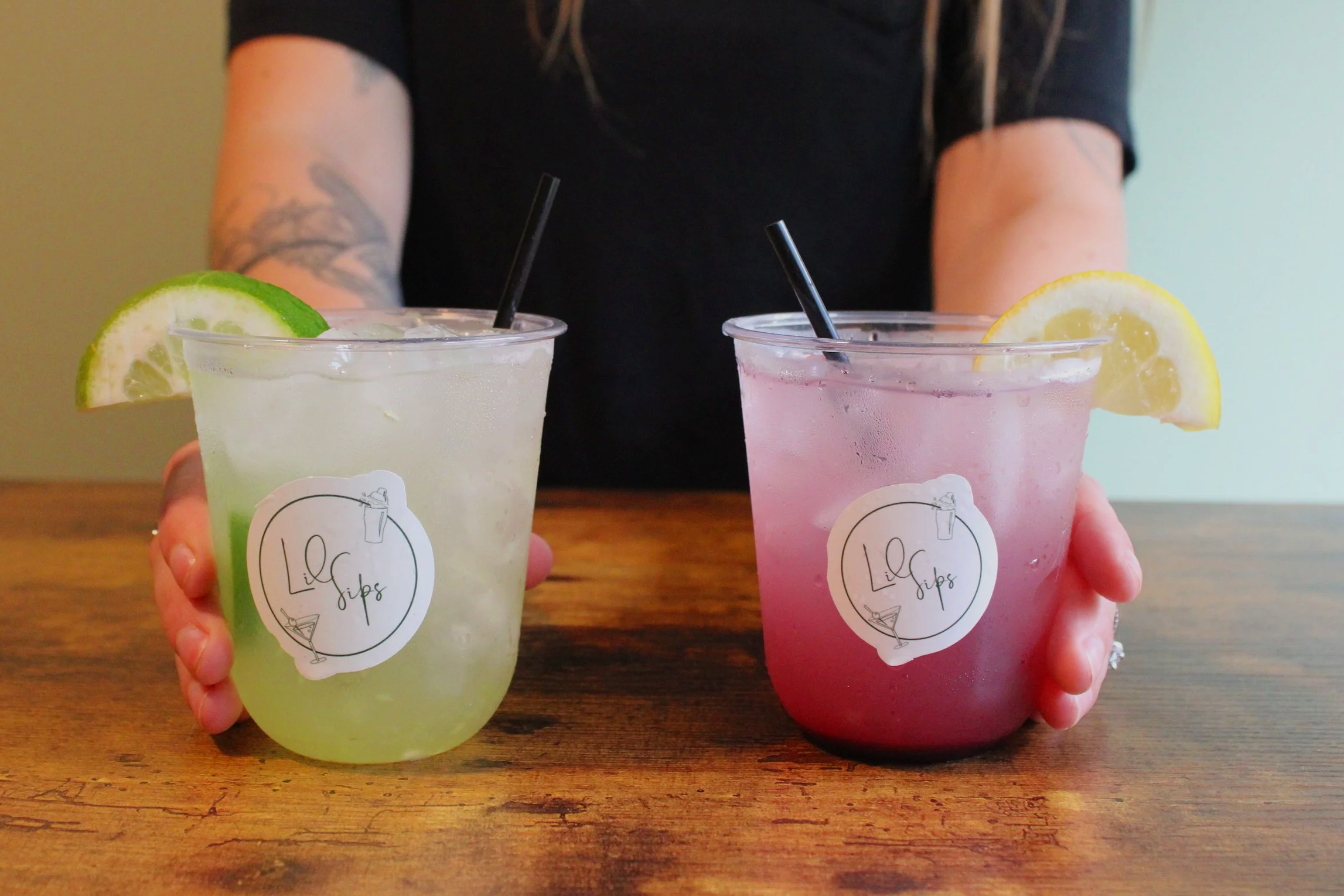 Two hands holding colorful drinks in clear cups with black straws. The left drink is pale yellow with a lime wedge, and the right drink is pink with a lemon wedge. Both cups have a white sticker with a logo for "LiliSips." A person with tattoos on their arms stands in the background against a neutral wall.