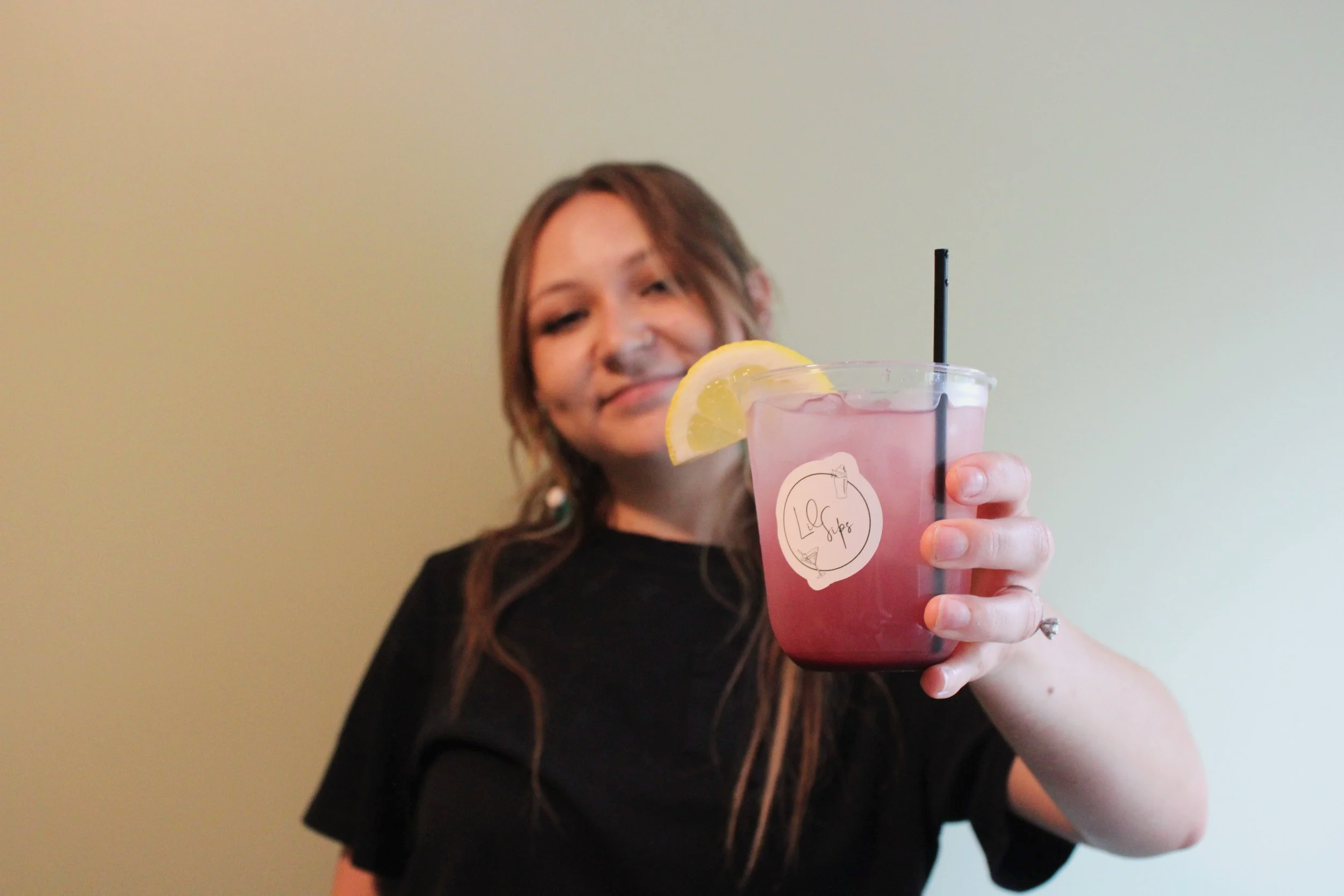 A woman with brown hair holding a pink fruit drink in a clear cup with a lemon slice and black straw, smiling.