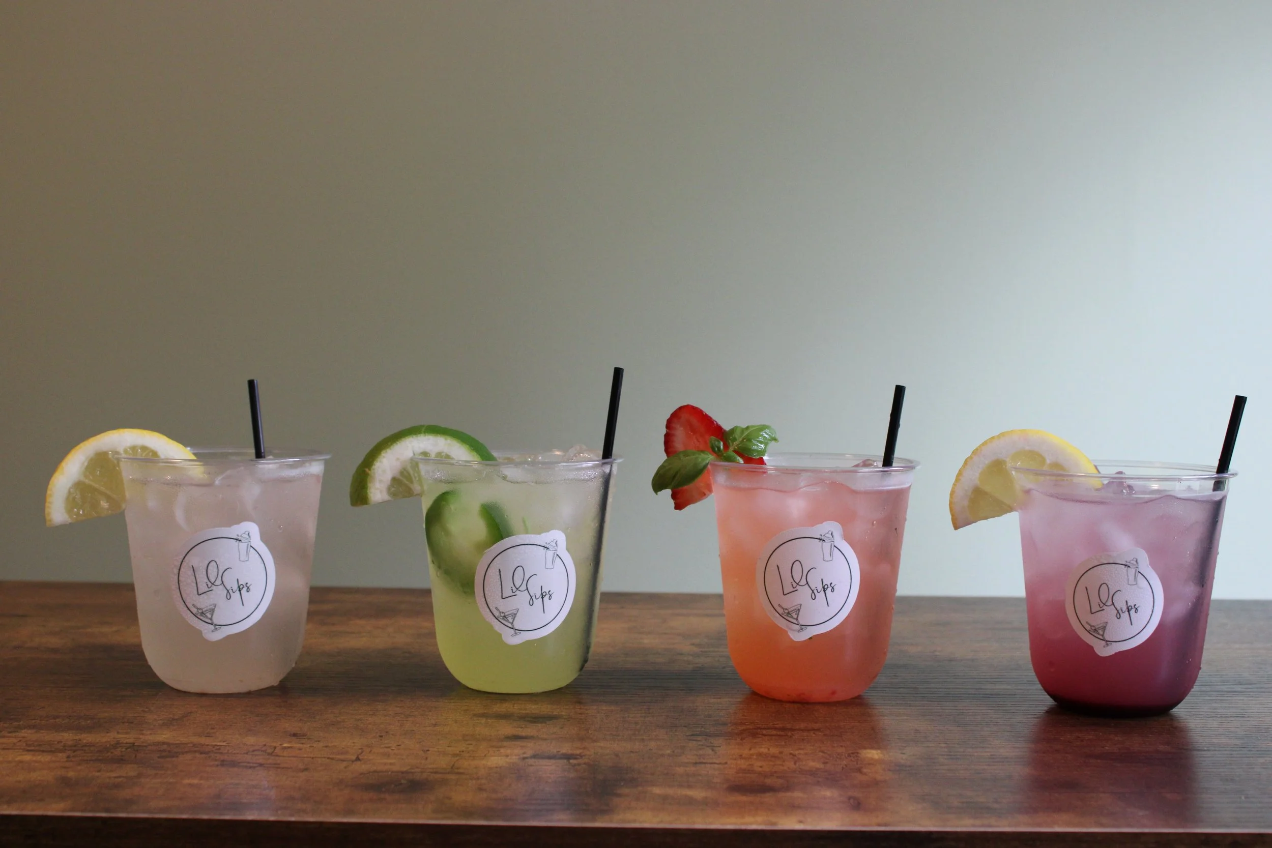 Four colorful beverages in clear cups with black straws, garnished with lemon, cucumber, strawberry, and lemon slices, on a wooden surface against a plain wall.
