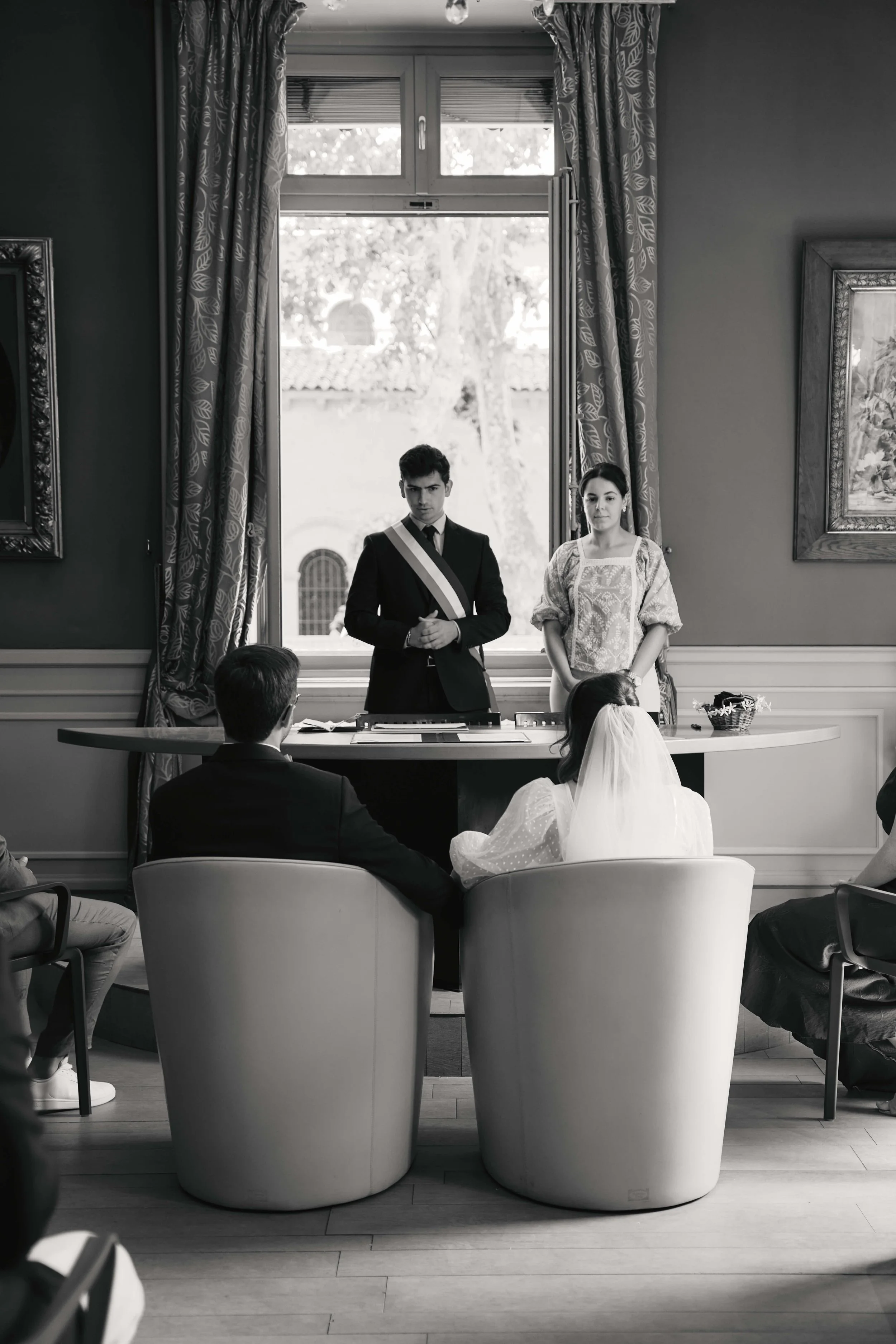A black and white photo of a room with a large window, two people standing in front of the window, and several people seated facing them, including a girl in a white dress and veil. The room has framed artwork on the walls and long curtains. The scen