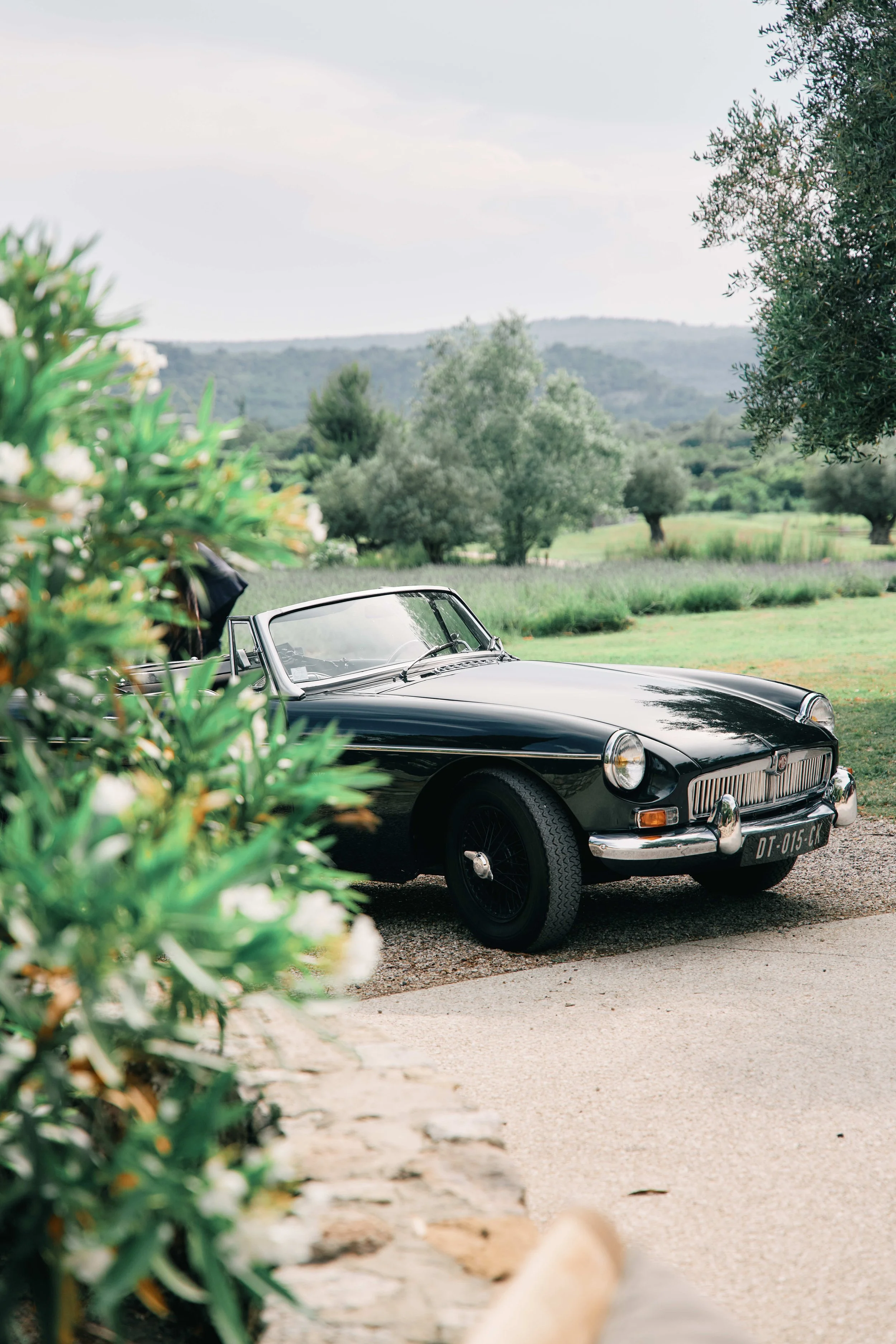 A vintage black convertible car parked on a gravel driveway in a lush green landscape with trees, hills, and cloudy sky in the background.