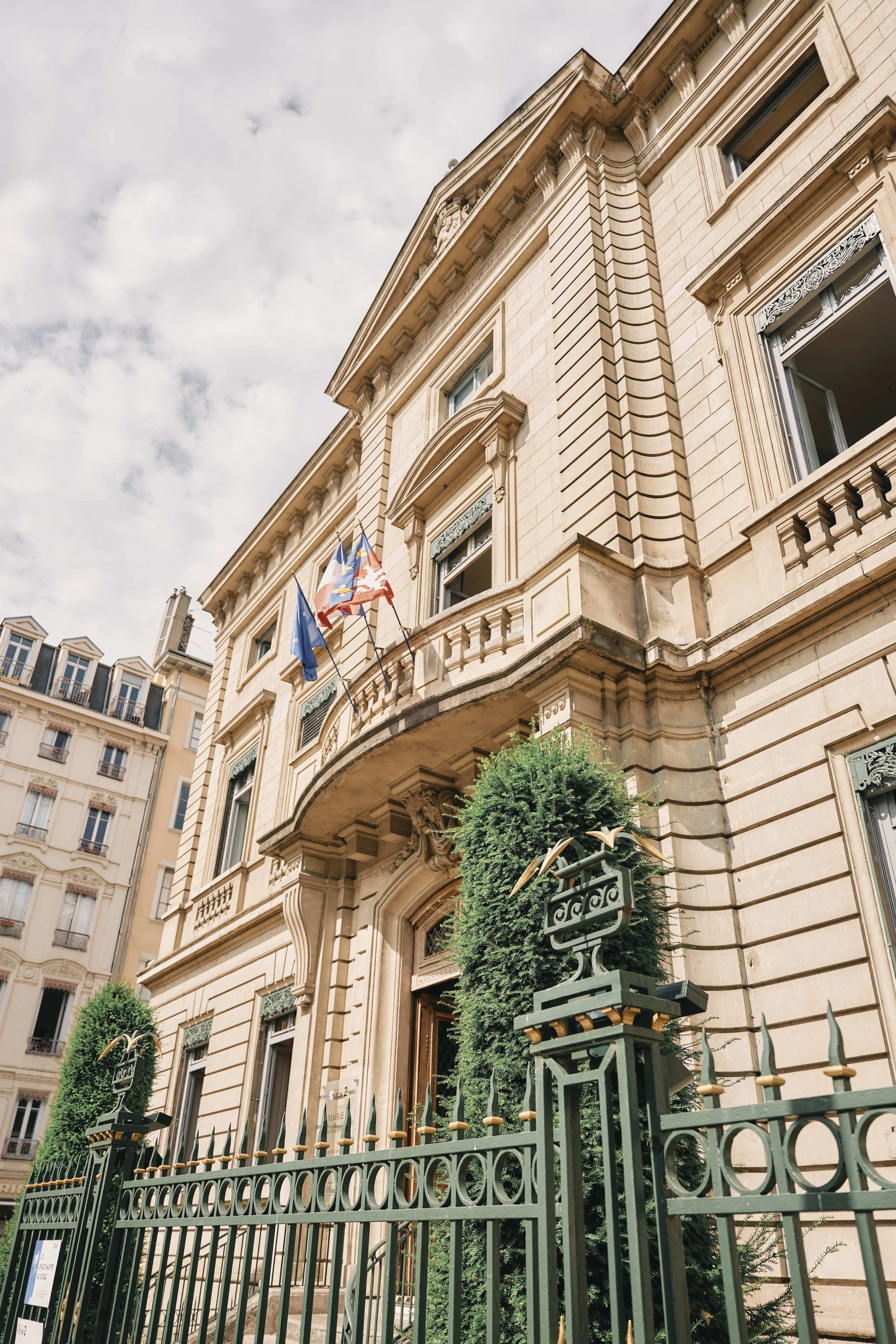 An ornate, beige stone building with tall windows and a balcony, decorated with flags and surrounded by a black wrought iron fence with gold accents, under a cloudy sky.