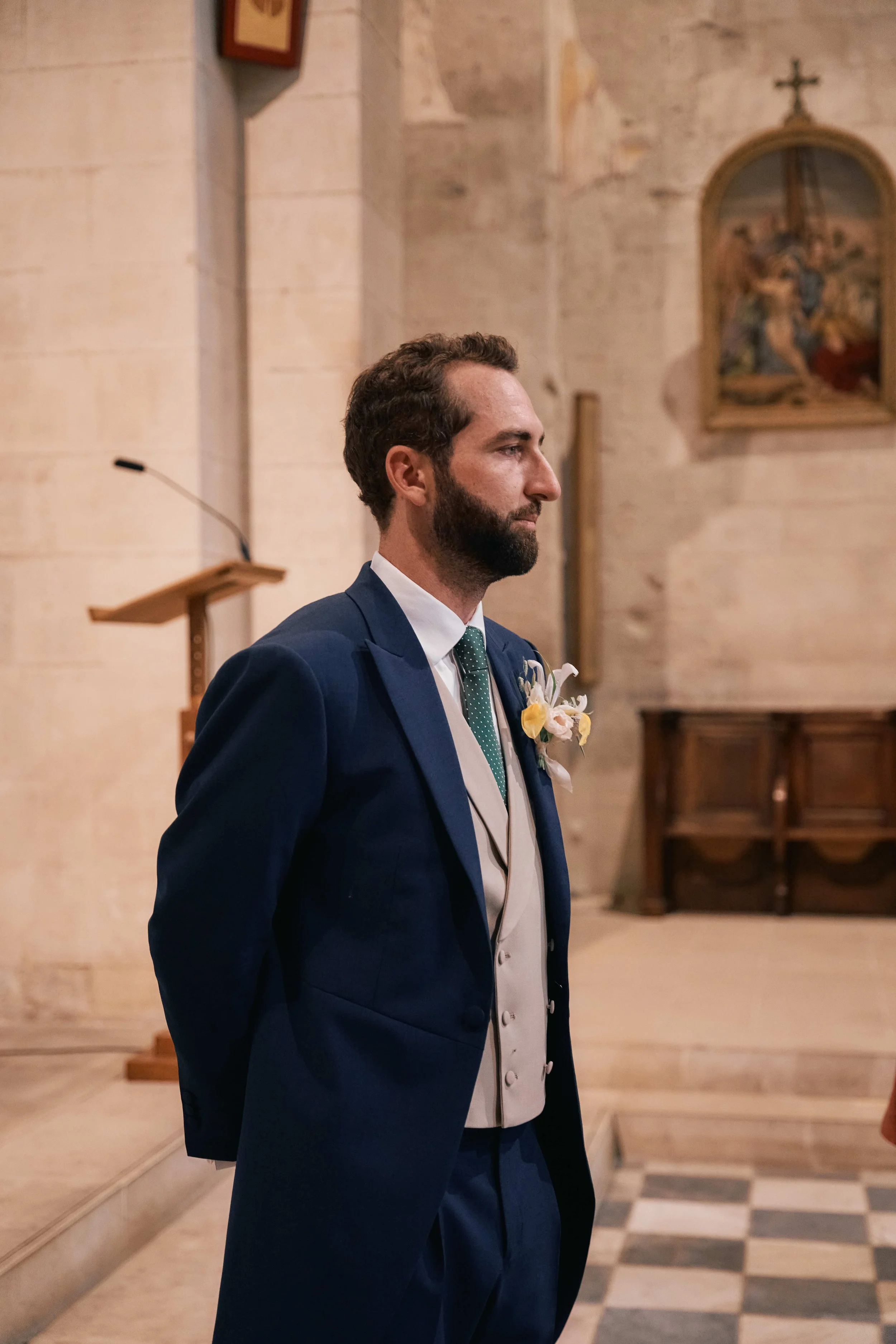 A man dressed in a dark blue suit with a white vest, wearing a green dotted tie and a boutonniere, standing inside a church or chapel with stone walls and religious paintings.