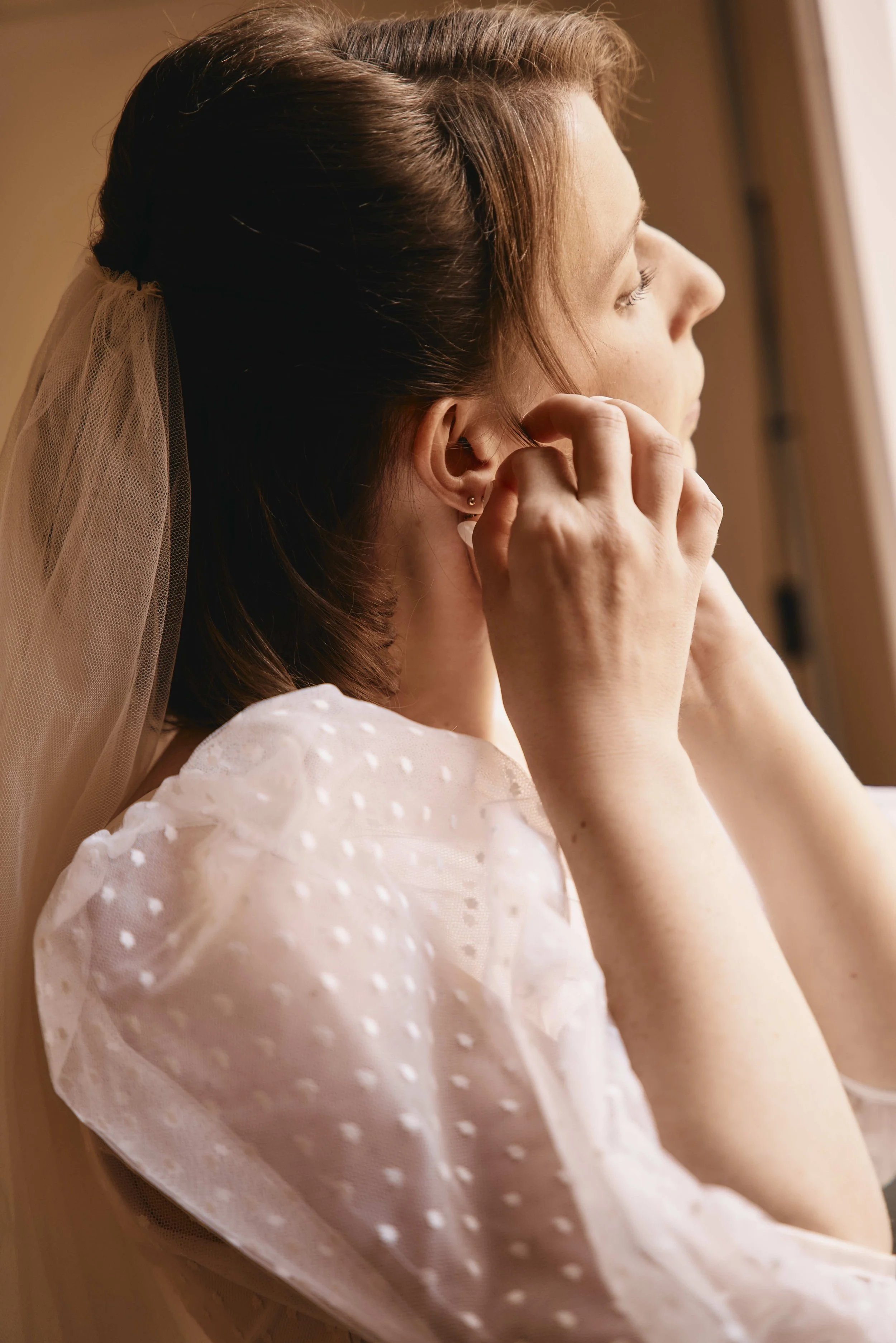 A woman with brown hair, wearing a veil, looking out a window, touching her ear, dressed in a white dress with polka dots.