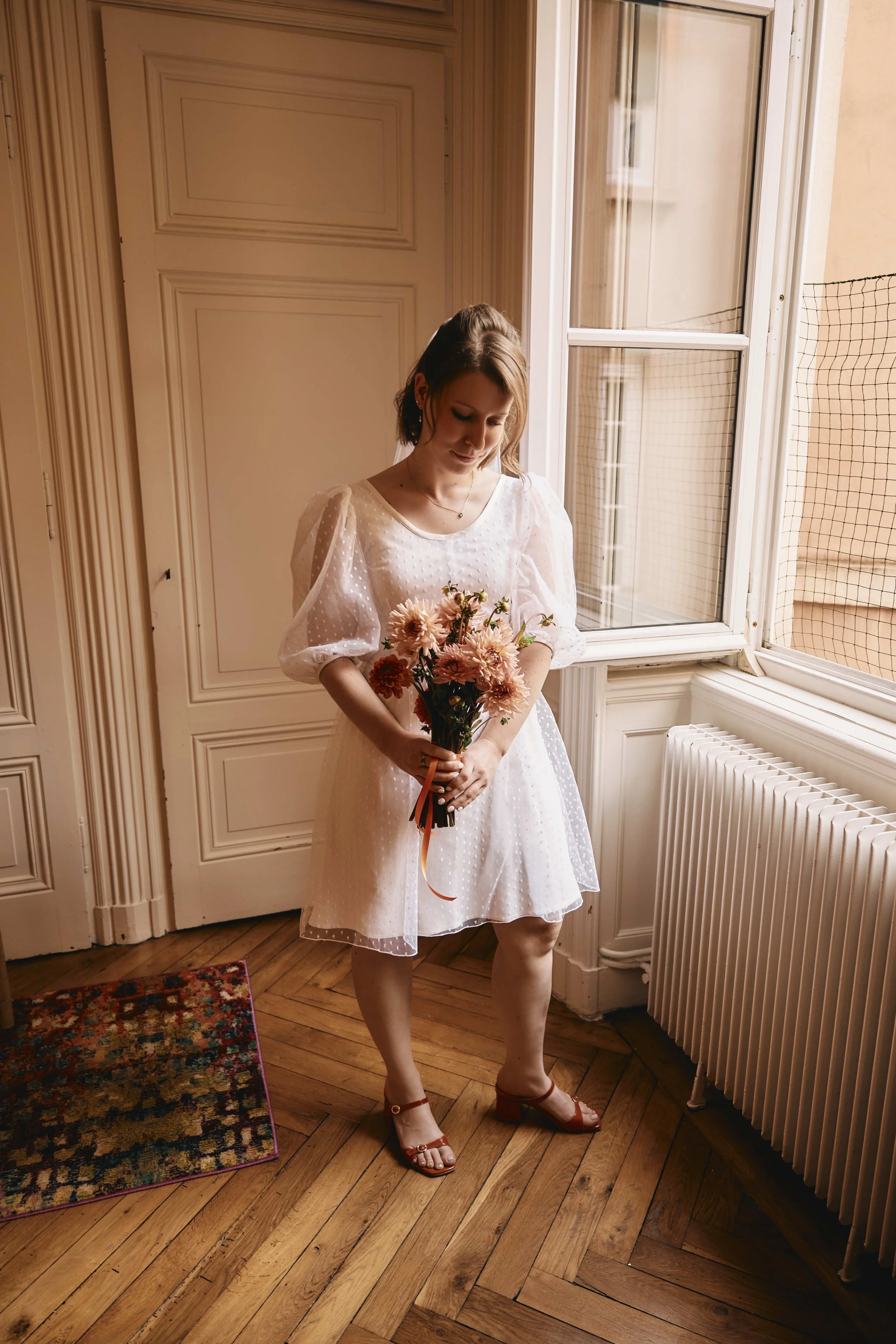 A woman in a white dress holding a bouquet of flowers standing by a window in a room with wooden flooring and paneled beige walls, wearing red sandals.
