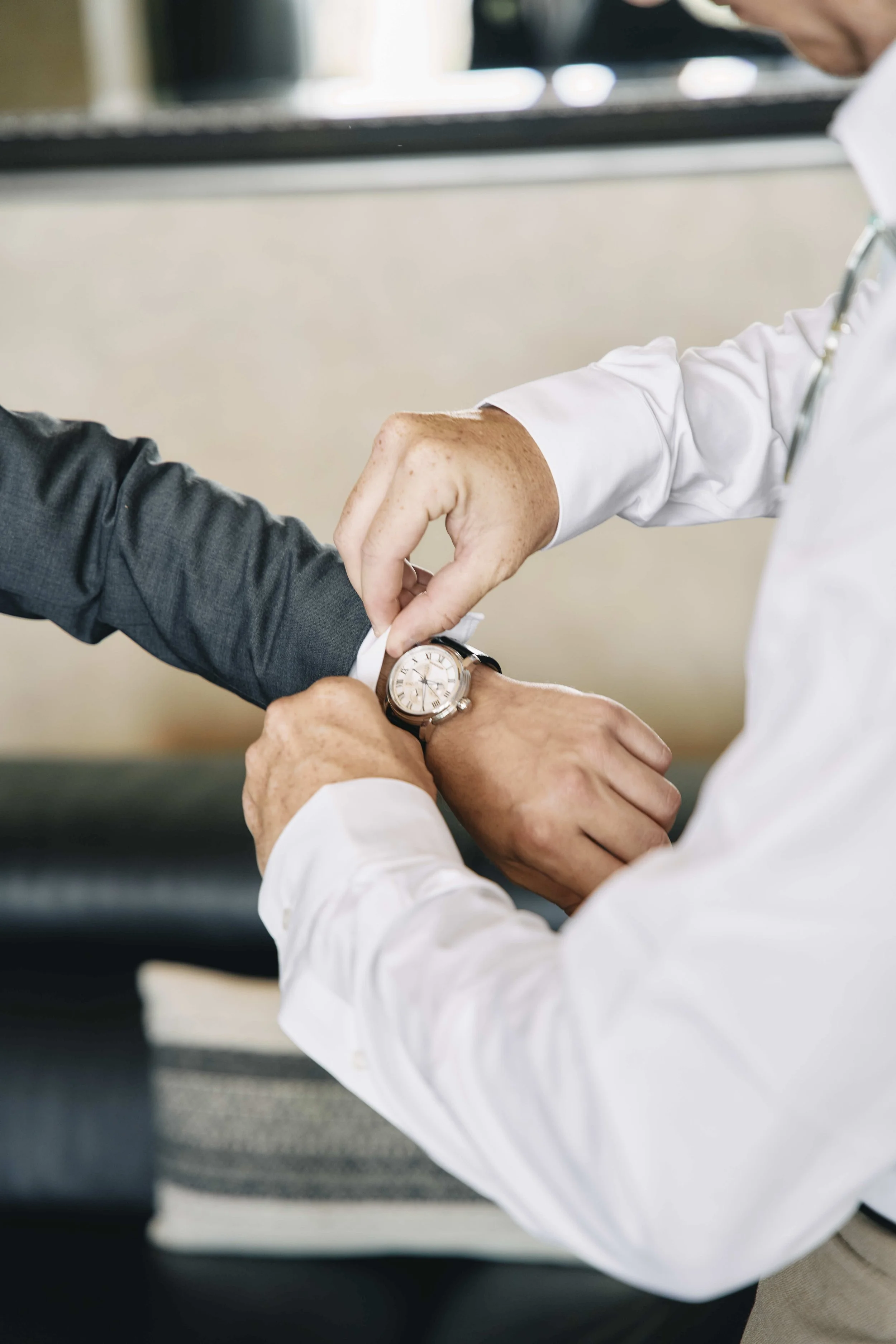 Two men, one wearing a gray jacket and the other in a white shirt, are adjusting or exchanging watches, with one adjusting the other's watch on his wrist.