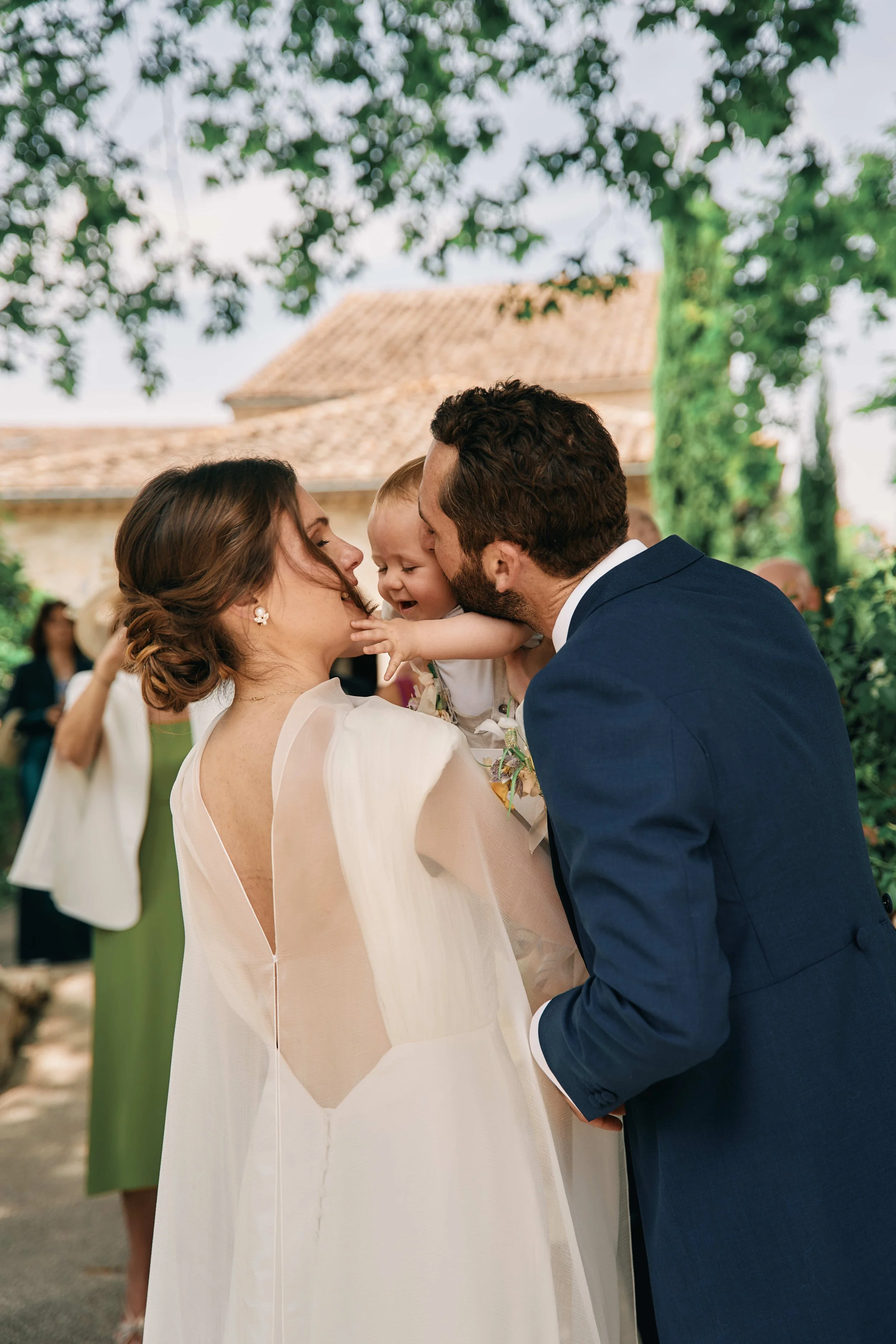 A smiling bride and groom are holding a young child at an outdoor wedding ceremony, with greenery and a building in the background.