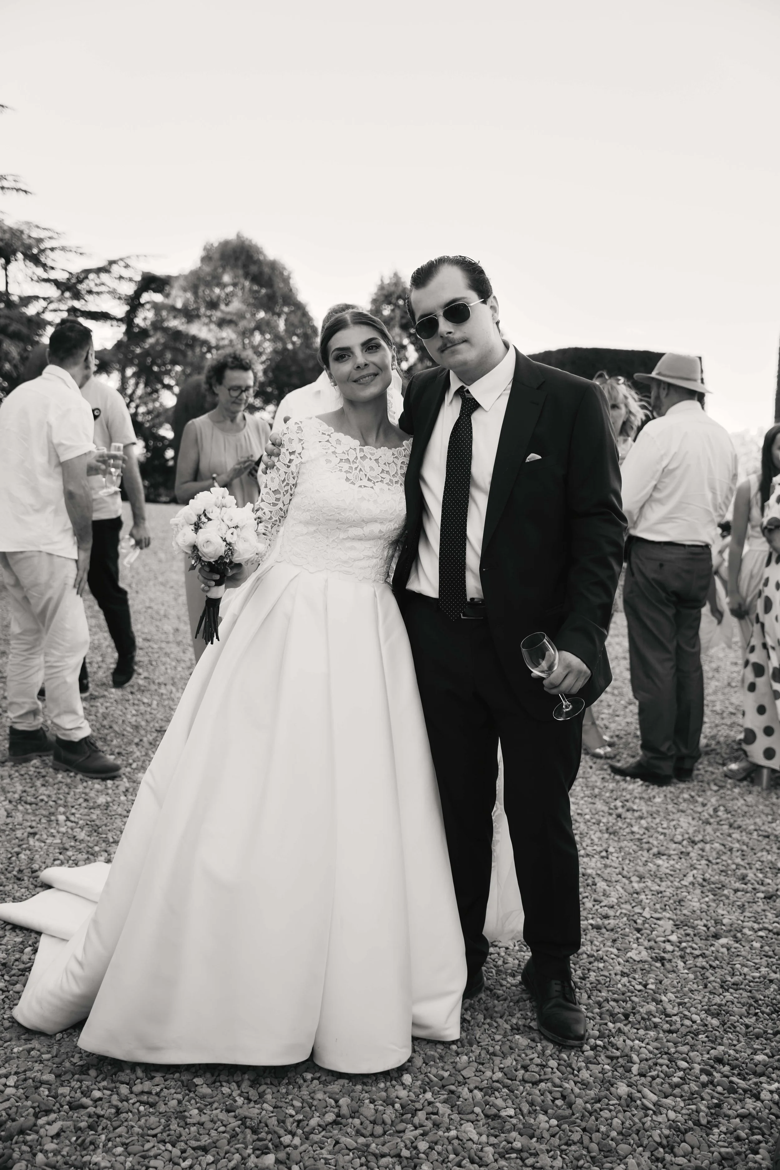 Bride in a white wedding gown holding a bouquet standing next to a groom in a black suit with sunglasses at an outdoor wedding reception.