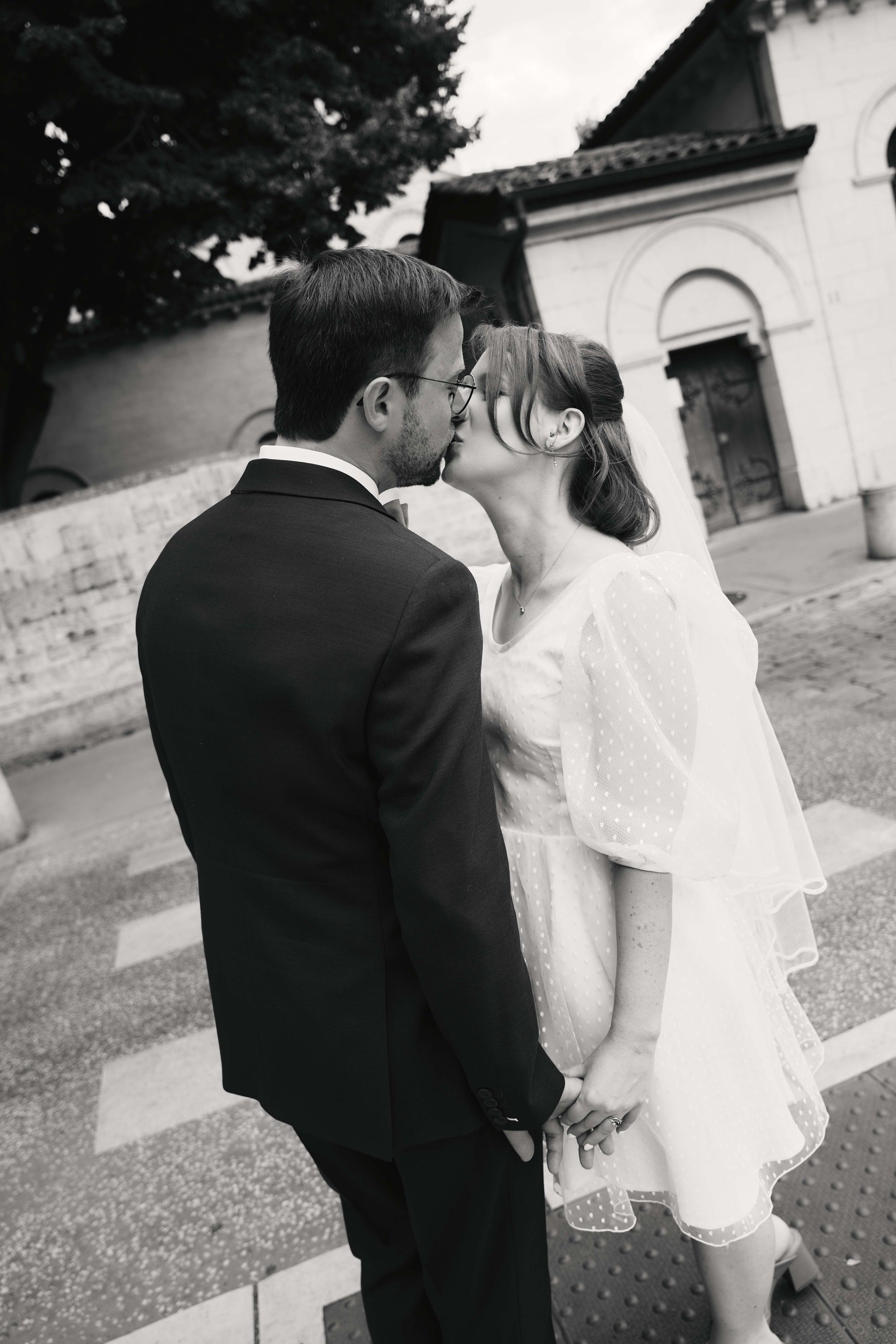 A black-and-white photograph of a bride and groom sharing a kiss outside a church. The groom is dressed in a black suit with a bow tie, and the bride is wearing a white dress with puffed sleeves and a veil. They are holding hands, and the bride's fac