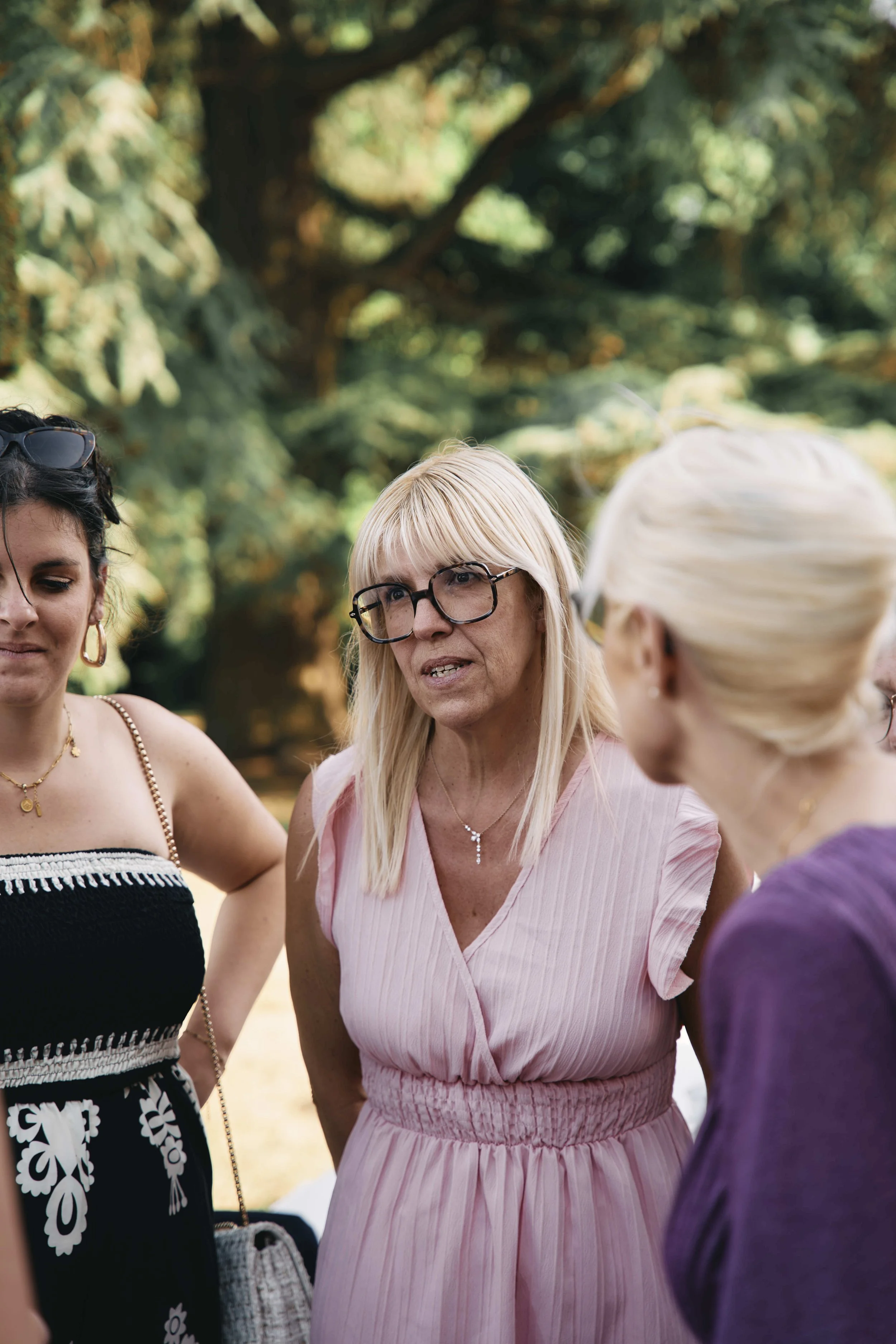 Four women standing together outdoors, engaged in a conversation. One woman in the middle wears glasses and a pink dress, with blonde hair. The other women have dark and white hair, dressed casually. The background features trees and greenery.