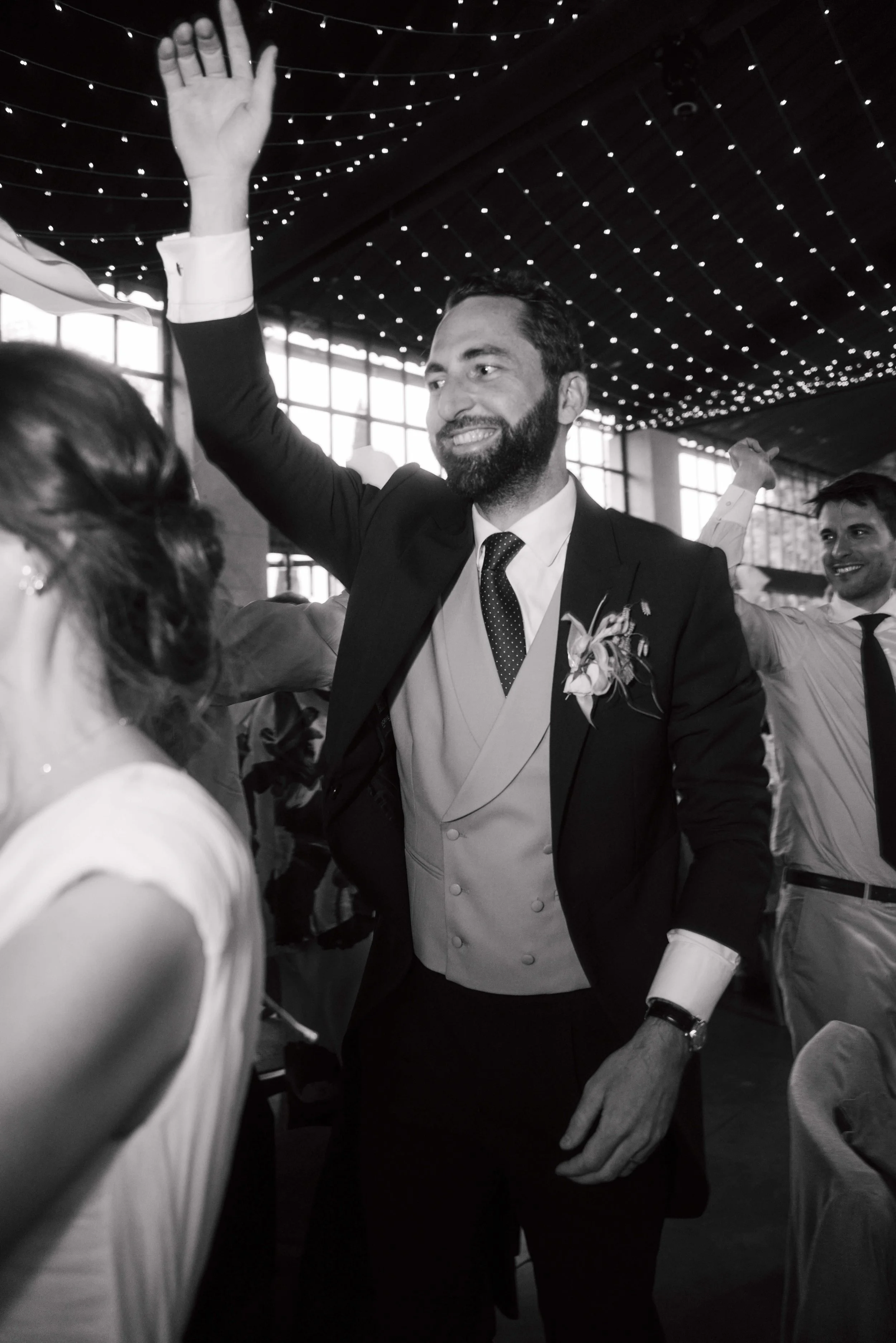 A man in a suit and tie at a wedding reception, raising his hand for a high-five with a woman. He has a boutonniere on his jacket. The background includes large windows, string lighting on the ceiling, and other guests smiling.