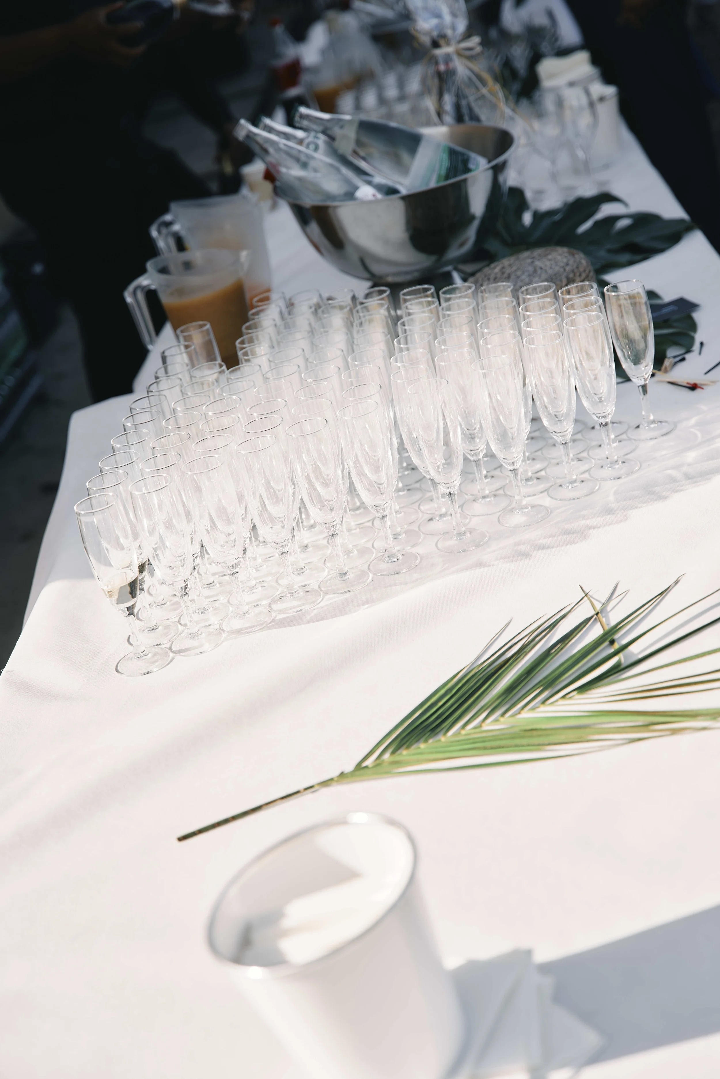 A table set for a celebration with rows of empty champagne glasses, a large silver bowl with bottles, and a green palm leaf on a white tablecloth.