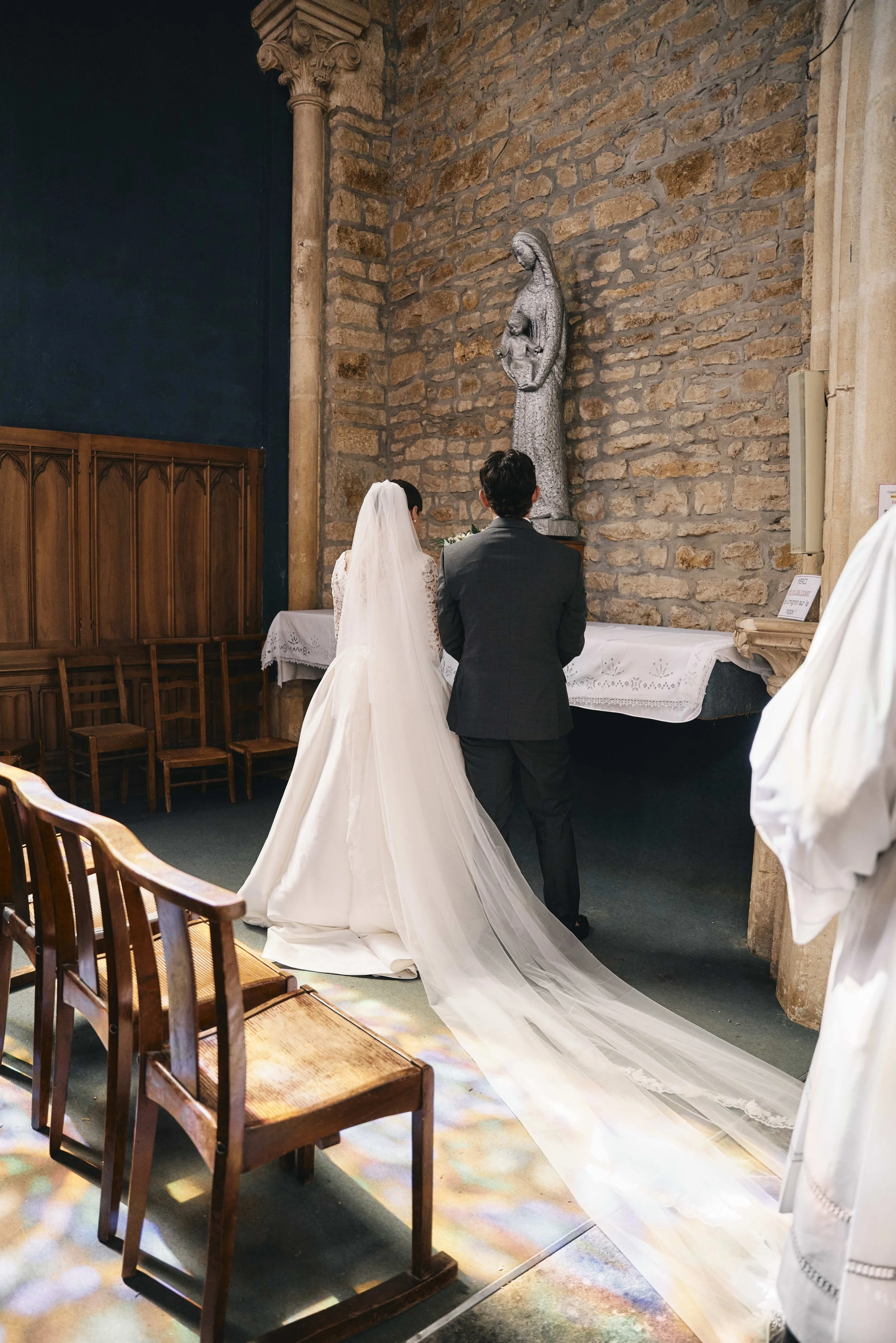 Bride and groom standing at the altar in a church with brick wall, a statue of Mary holding baby Jesus behind them, wedding altar covered with white cloth, wooden chairs on side, sunlight creating colorful reflections on the floor.