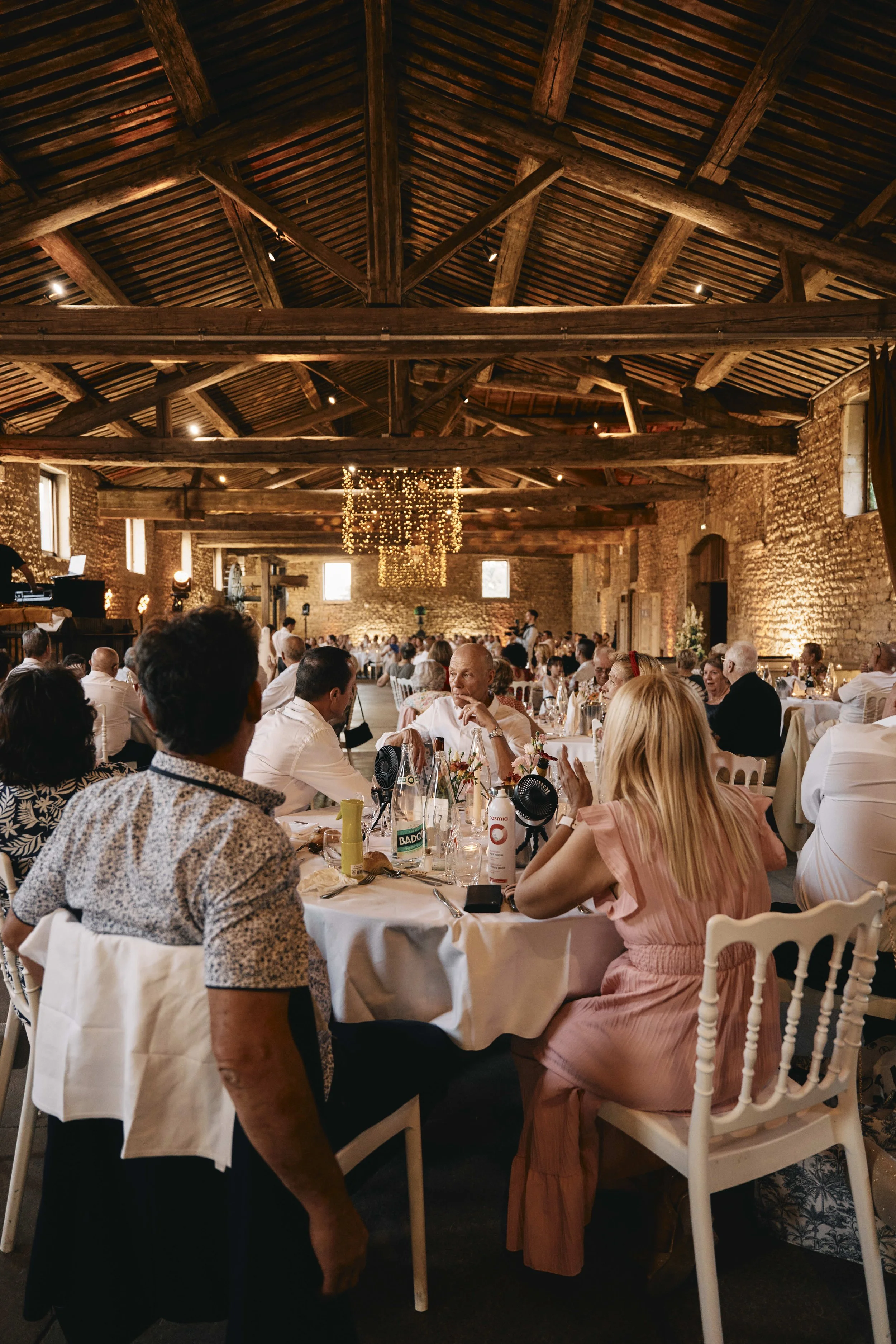 Interior of a rustic event space with exposed brick walls and wooden beams. Attendees seated at tables engaged in conversation, with warm lighting and string lights hanging from the ceiling.