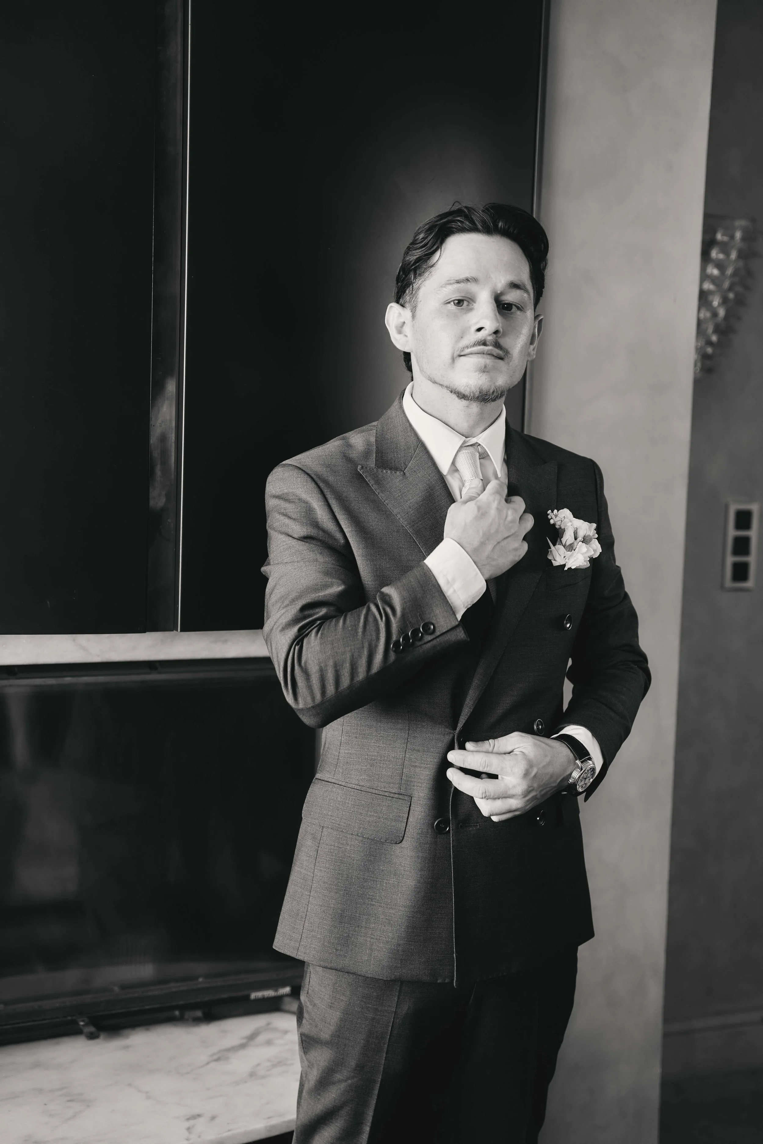 A man in a suit adjusting his tie on a marble surface, standing in front of a dark wall and elevator panel.