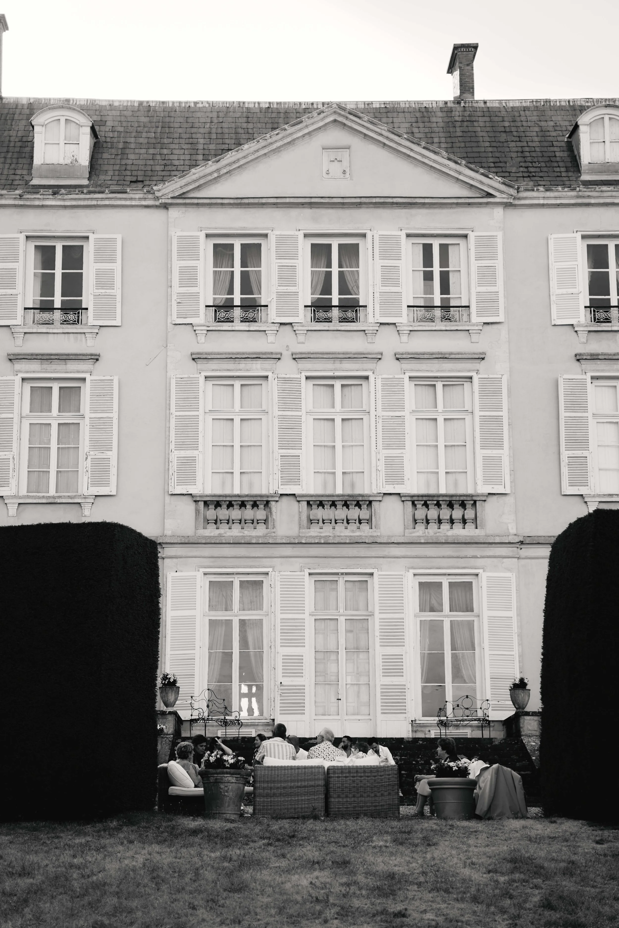 People dining outdoors in front of an elegant, multi-story building with tall windows and white shutters, in a garden setting with a well-manicured lawn and large potted plants.