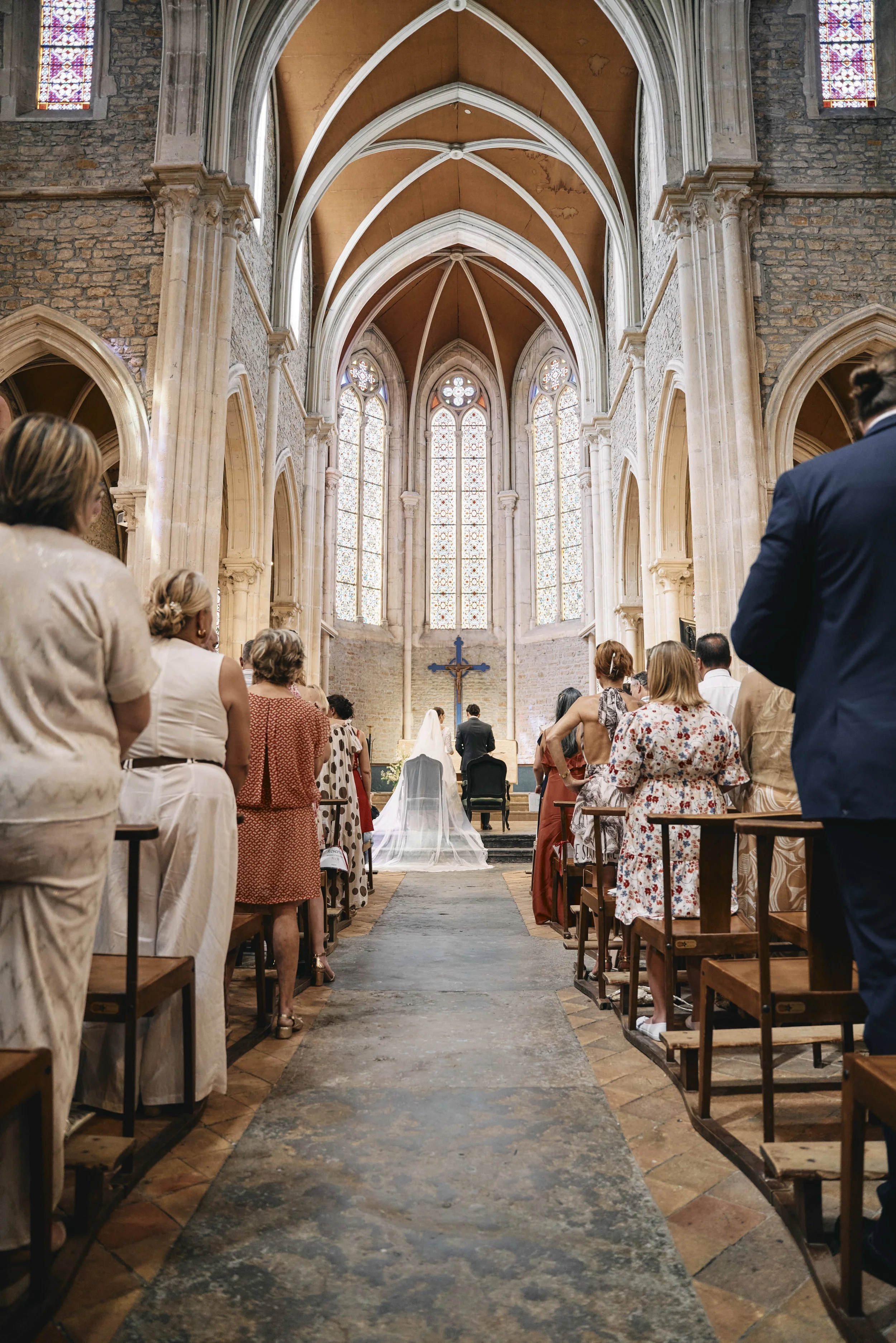 Inside a church during a wedding ceremony with people sitting and standing, an altar with a cross and wedding dress, large stained glass windows, stone walls, vaulted ceilings, and wooden pews.