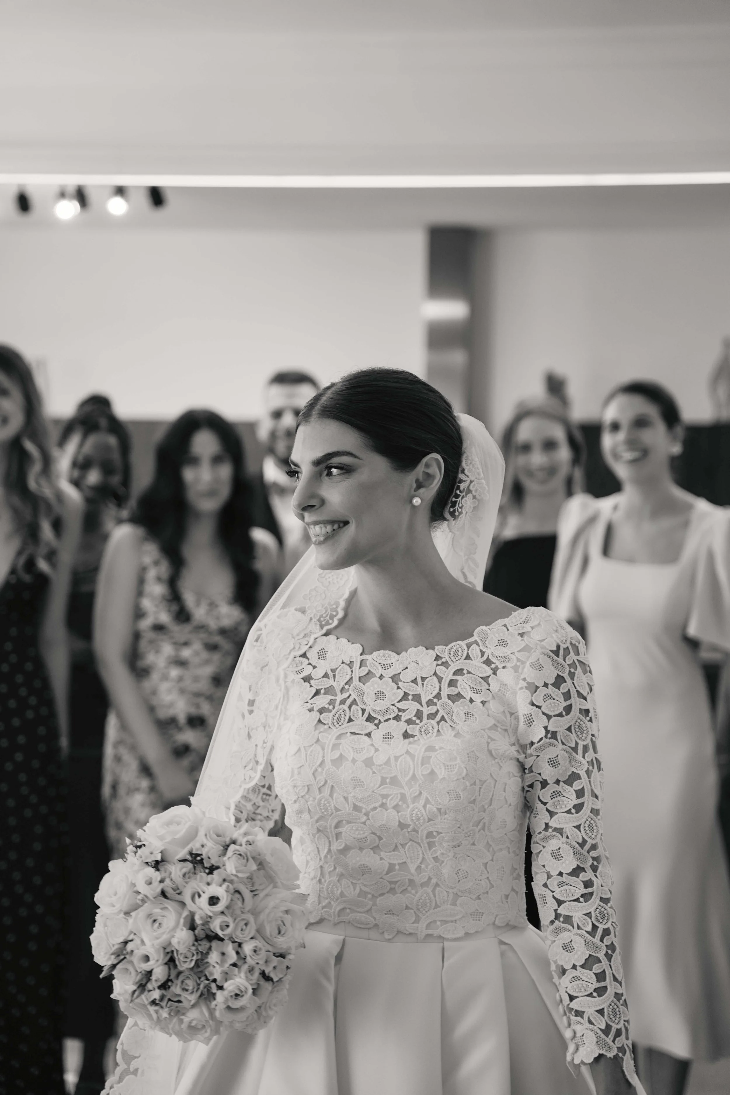 Black and white photo of a bride with dark hair in an elegant lace wedding dress holding a bouquet of roses, surrounded by women in formal attire smiling.