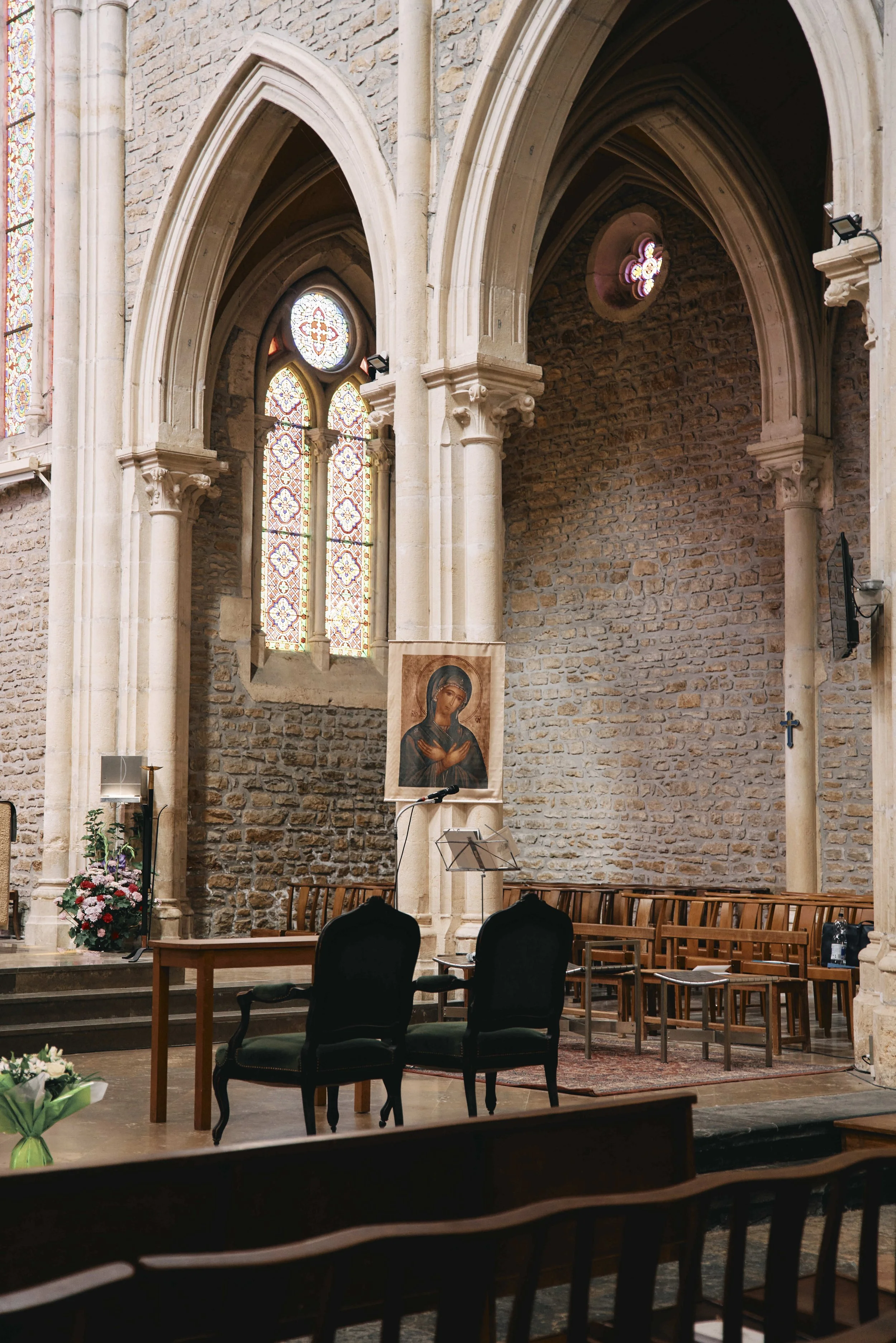 Interior of a church with stone walls, stained glass windows, wooden chairs, a table with a bouquet of flowers, and religious images.