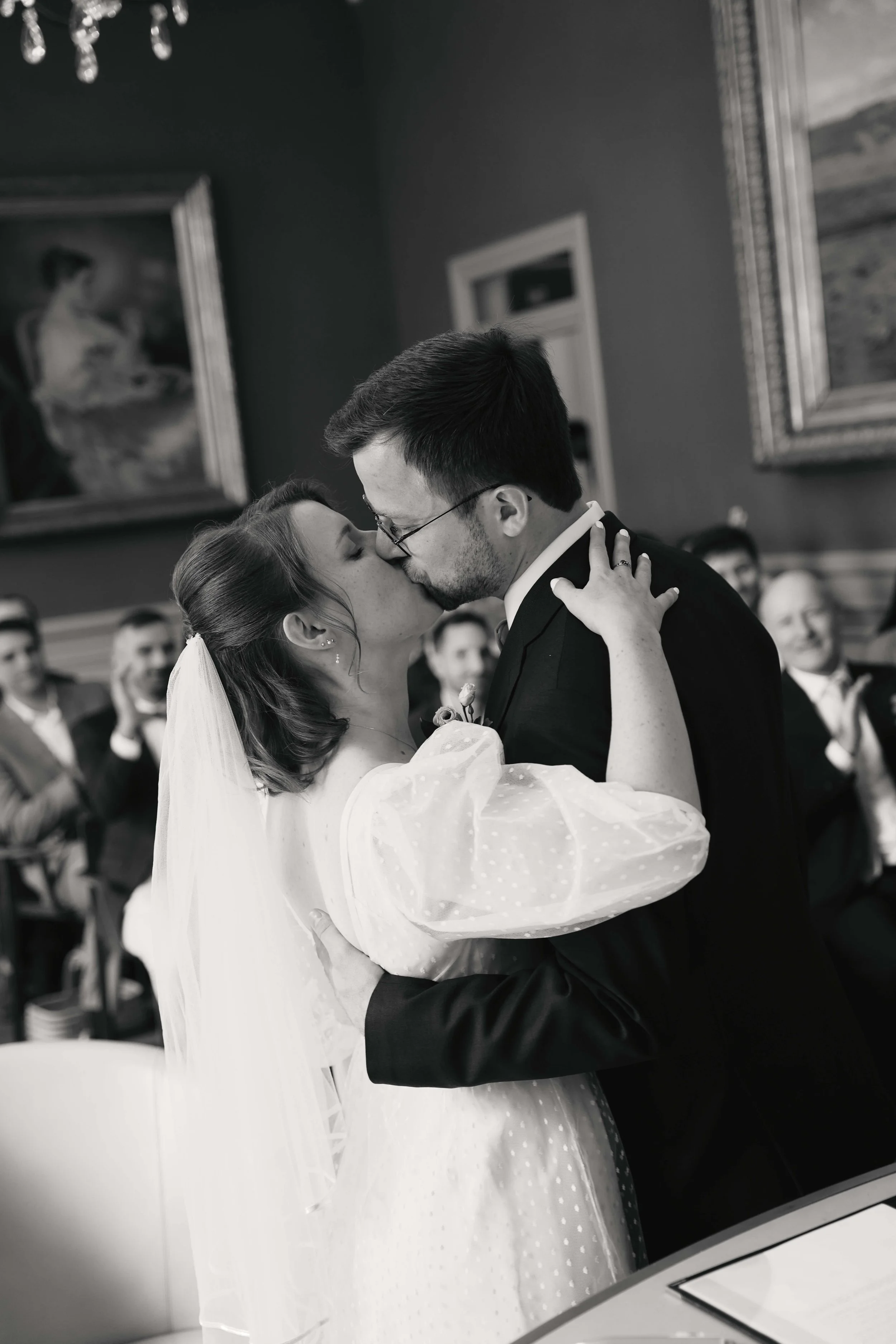 A bride and groom share a kiss during their wedding ceremony in a decorated indoor setting, with guests watching in the background.