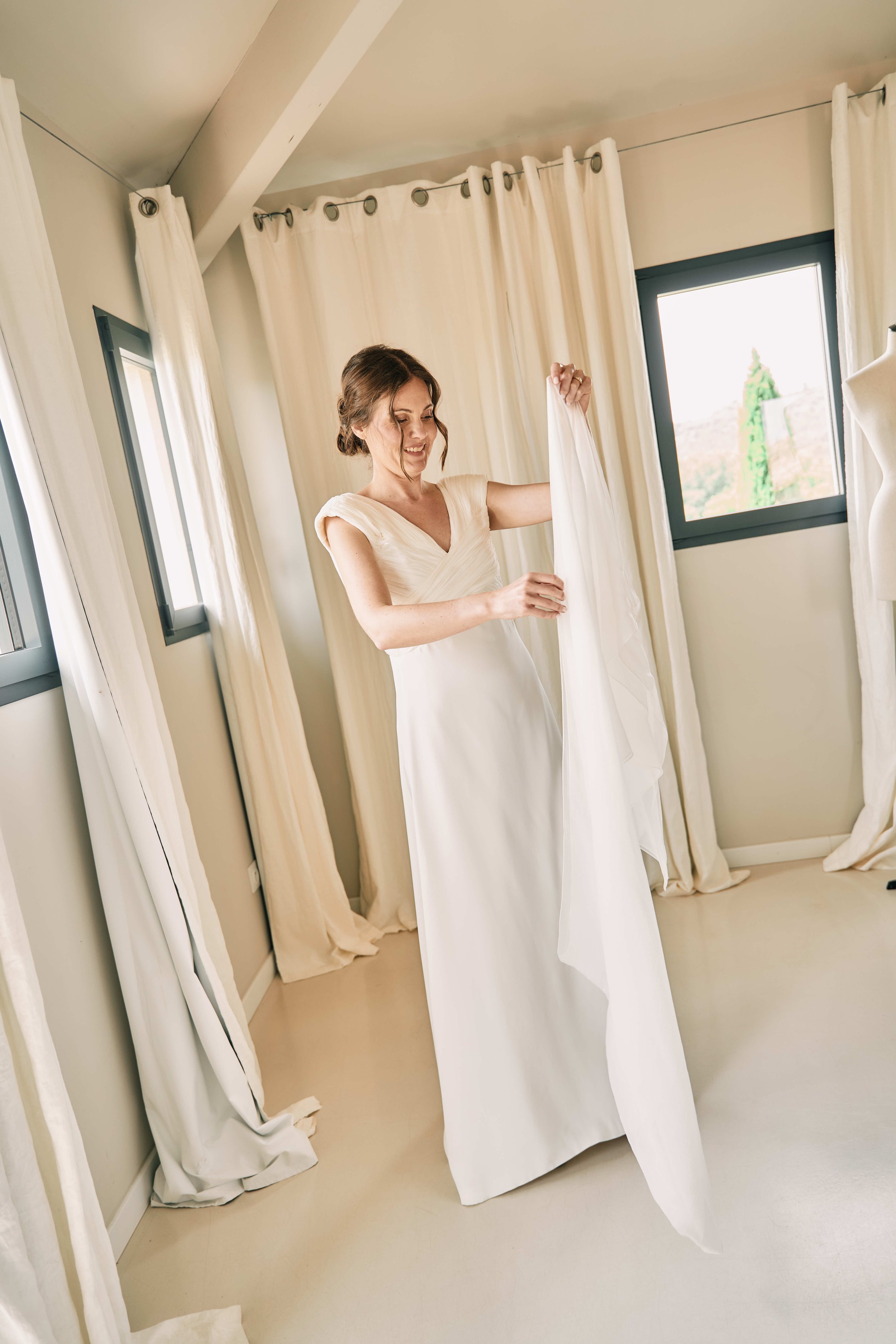 A woman in a white dress smiling as she touches the fabric of her wedding dress, standing in a bright room with cream-colored curtains and windows.