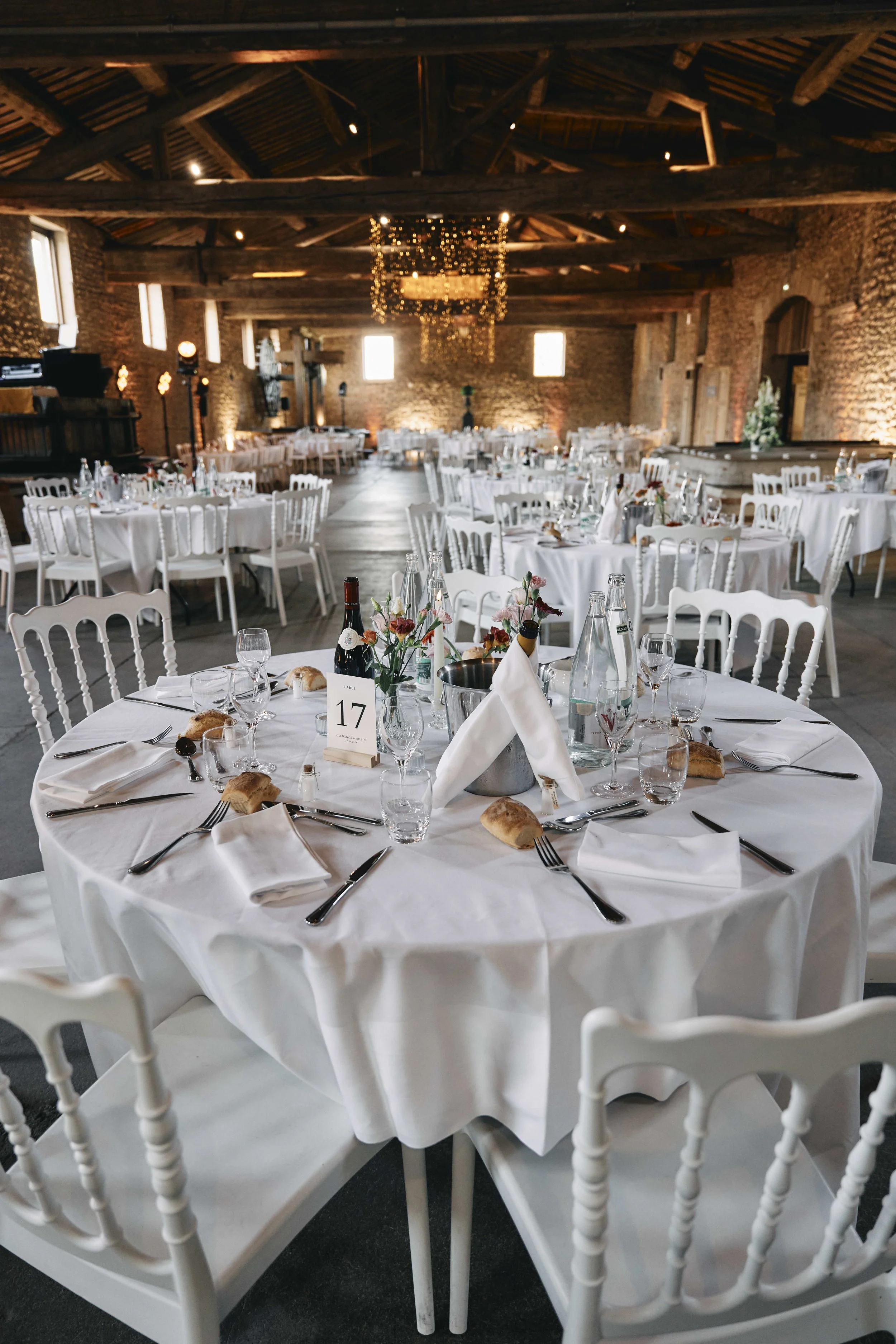 Wedding reception setup inside a rustic barn with round tables covered in white tablecloths, decorated with flowers, candles, and place settings, featuring white wooden chairs and a chandelier with string lights hanging from the wooden ceiling.