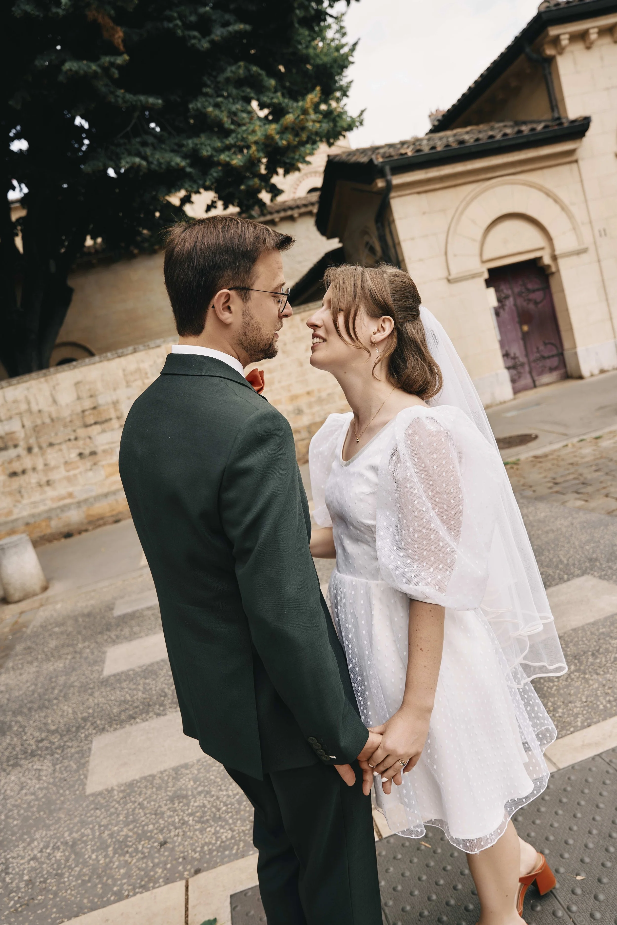 A newlywed couple holding hands and gazing at each other outdoors, near a historic stone building with arched doorways, a large tree, and a cobblestone sidewalk.