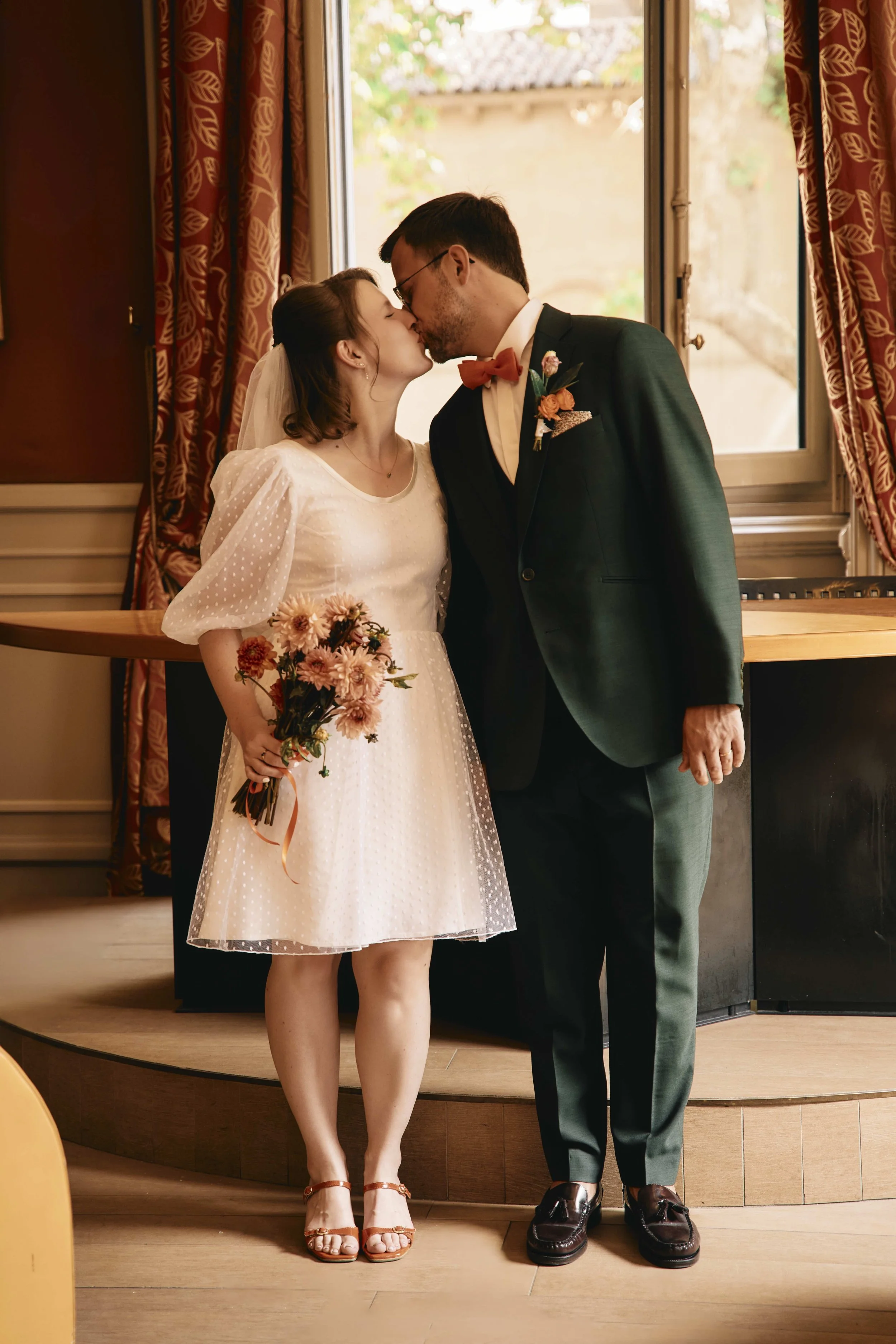 A bride and groom sharing a kiss indoors. The bride is wearing a white dotted dress, a veil, and tan high-heeled sandals. She is holding a bouquet of pink and orange flowers. The groom is in a dark suit, white shirt, red bow tie, and black shoes. The