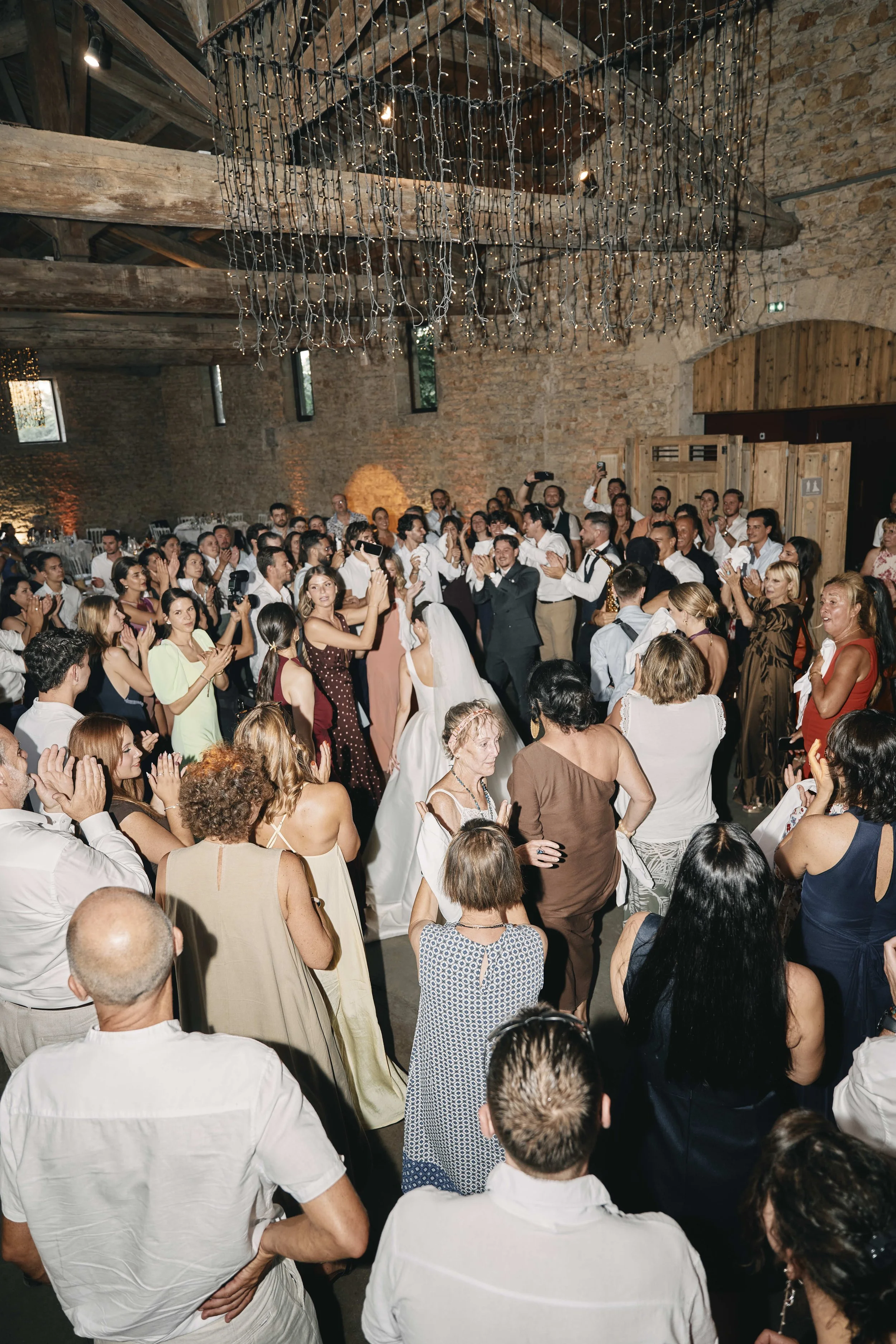 A lively indoor wedding reception with guests dancing and celebrating in a rustic barn with wooden beams and stone walls, decorated with string lights.