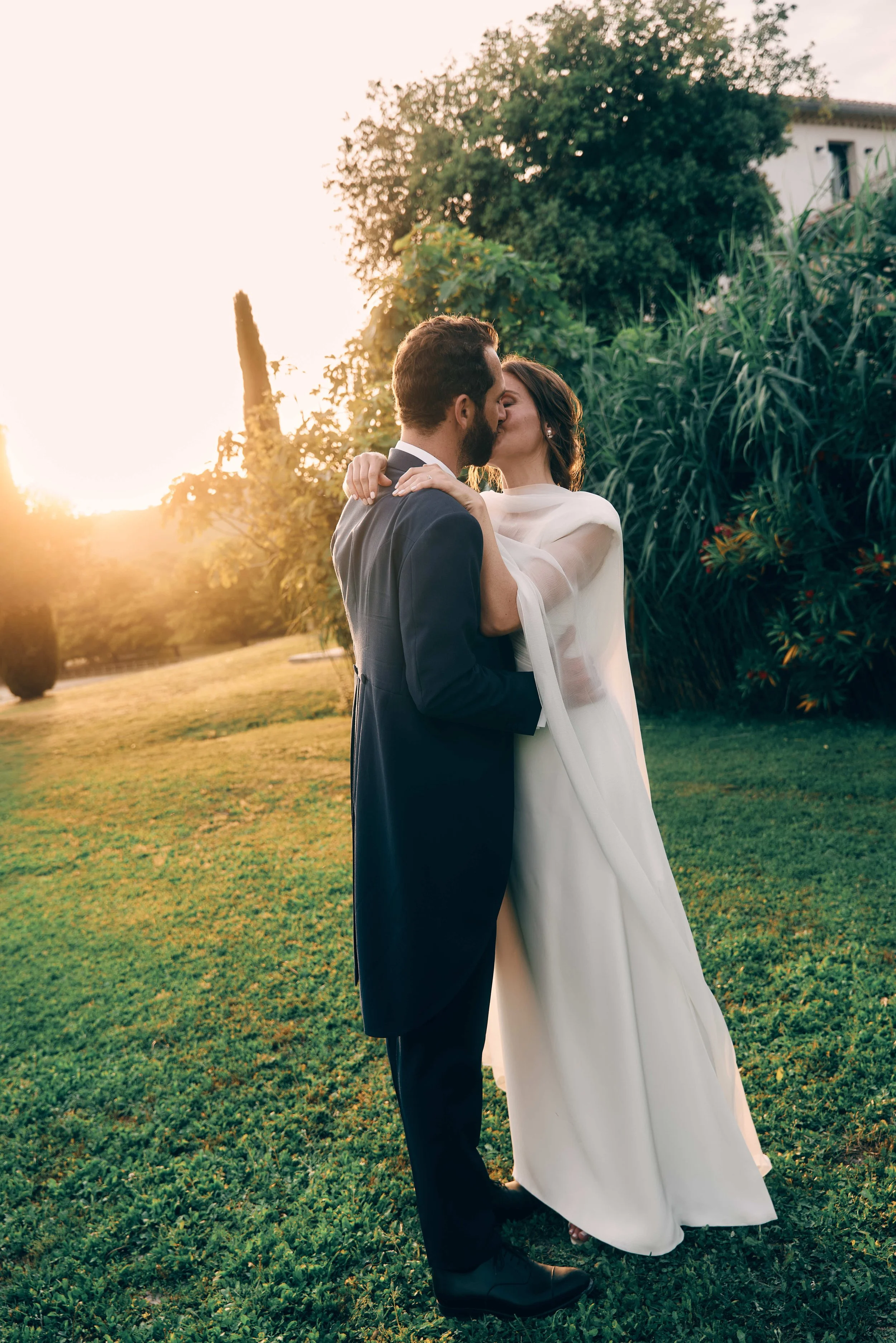 A newlywed couple sharing a kiss outdoors during sunset, with greenery and tall trees in the background.
