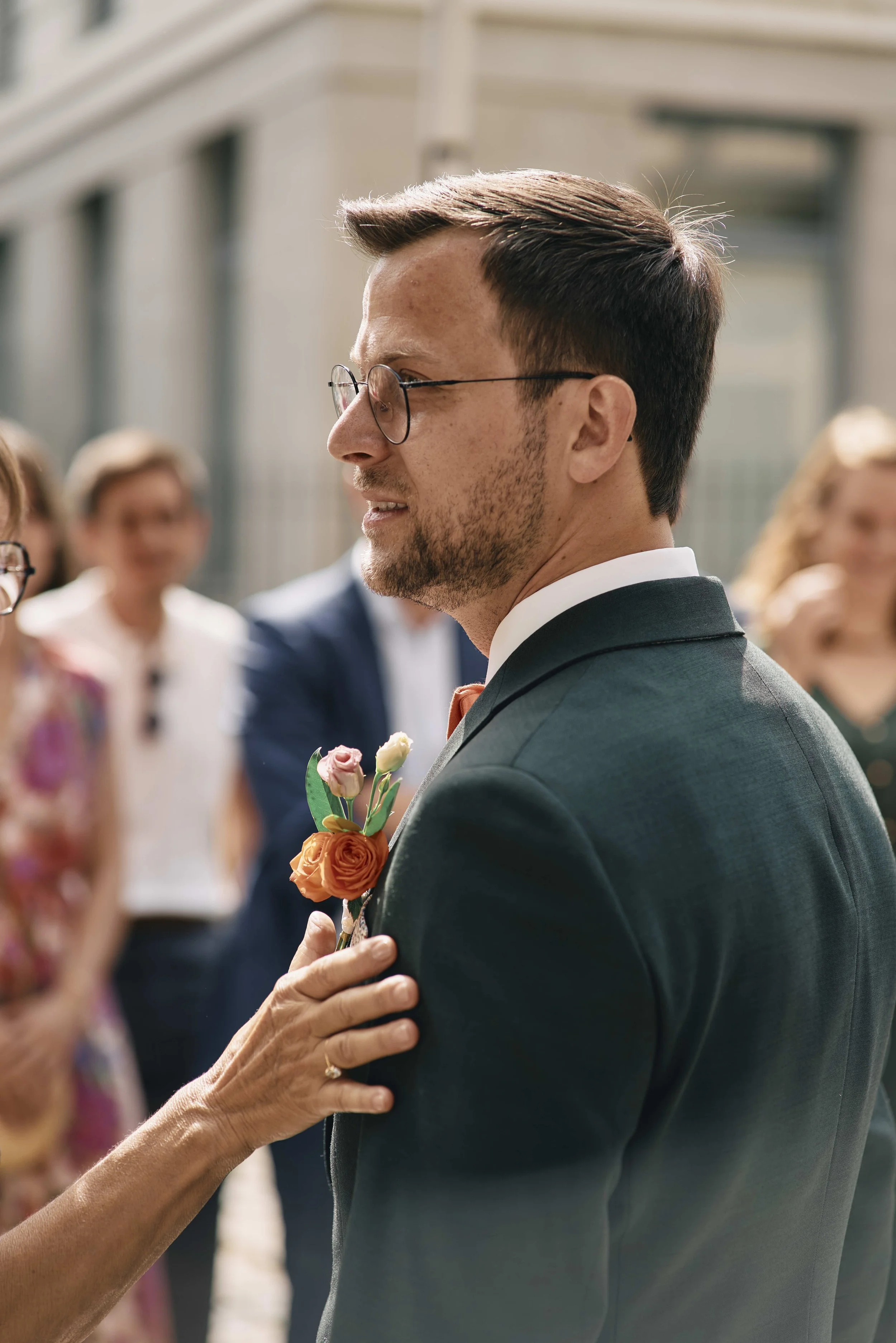 A young man with glasses in a suit at a wedding, with a woman reaching towards him. The woman holds a small bouquet with orange and pink roses.