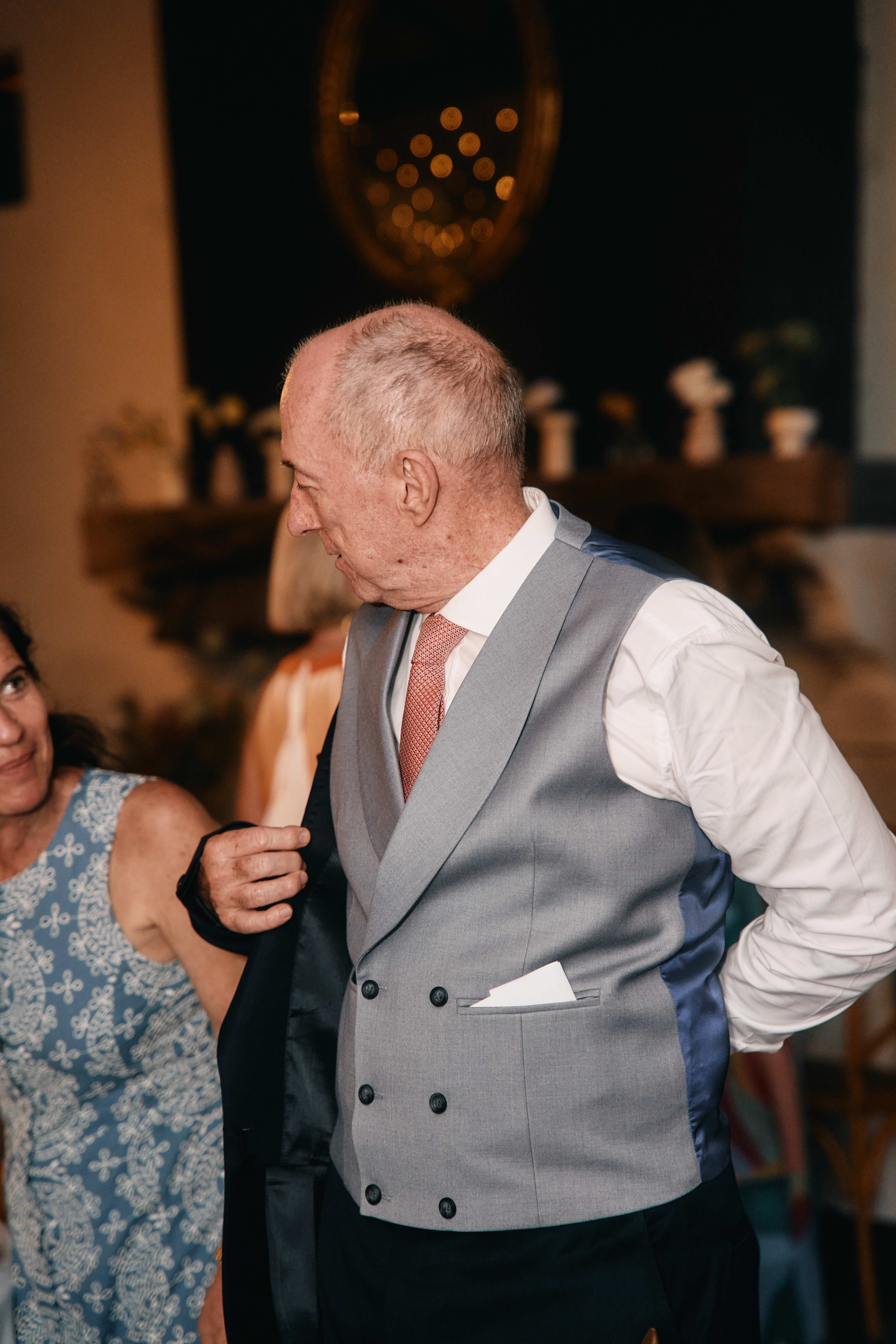 A man in a light gray vest, white shirt, and pink tie is dancing with a woman who is wearing a blue dress with white floral patterns. The background shows a dark wall with a decorative mirror and shelves with small potted plants. The man has a slight