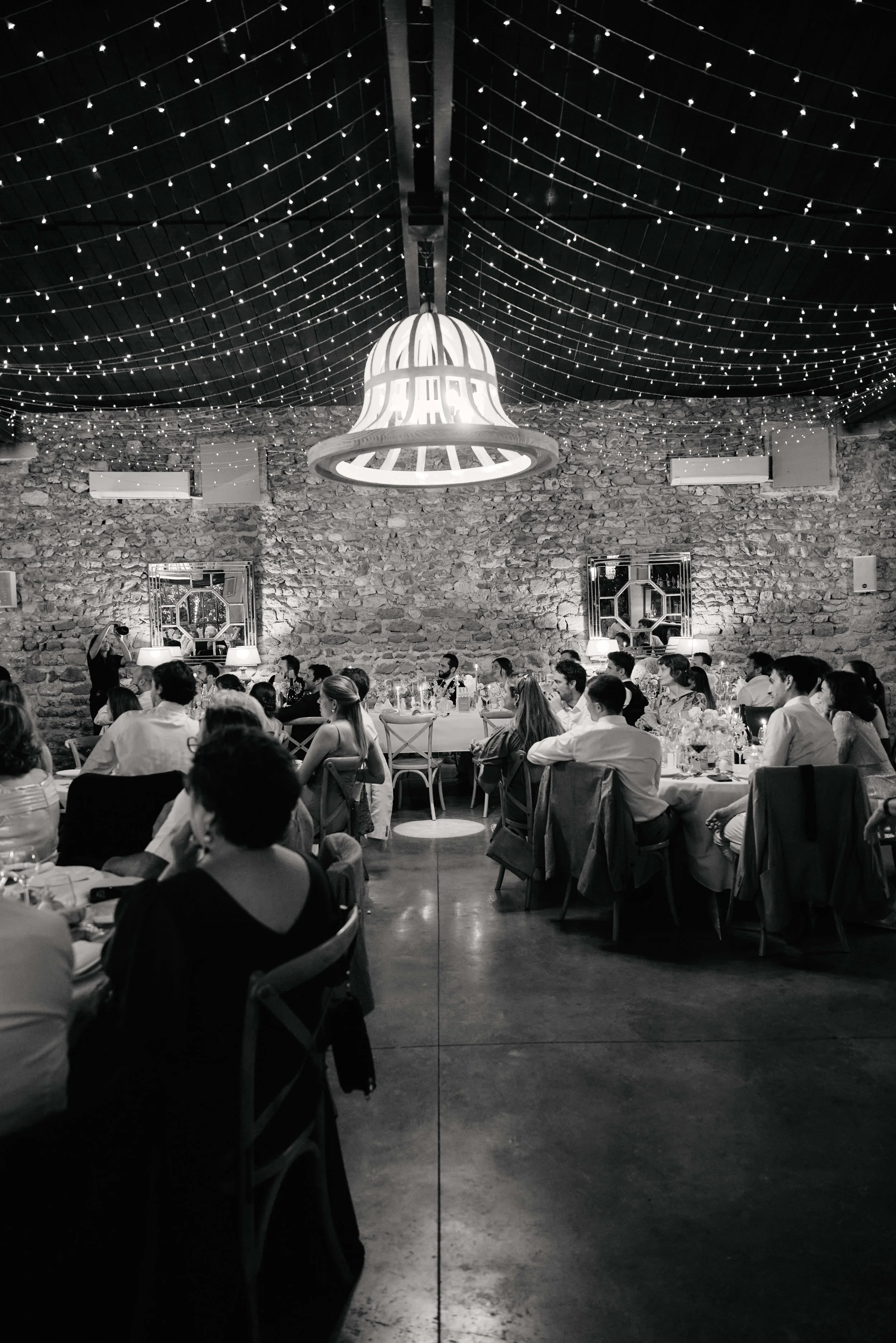A large gathering of people seated at round tables in a rustic event space with stone walls and string lights hanging from the ceiling. A large chandelier or decorative light fixture hangs above the room.