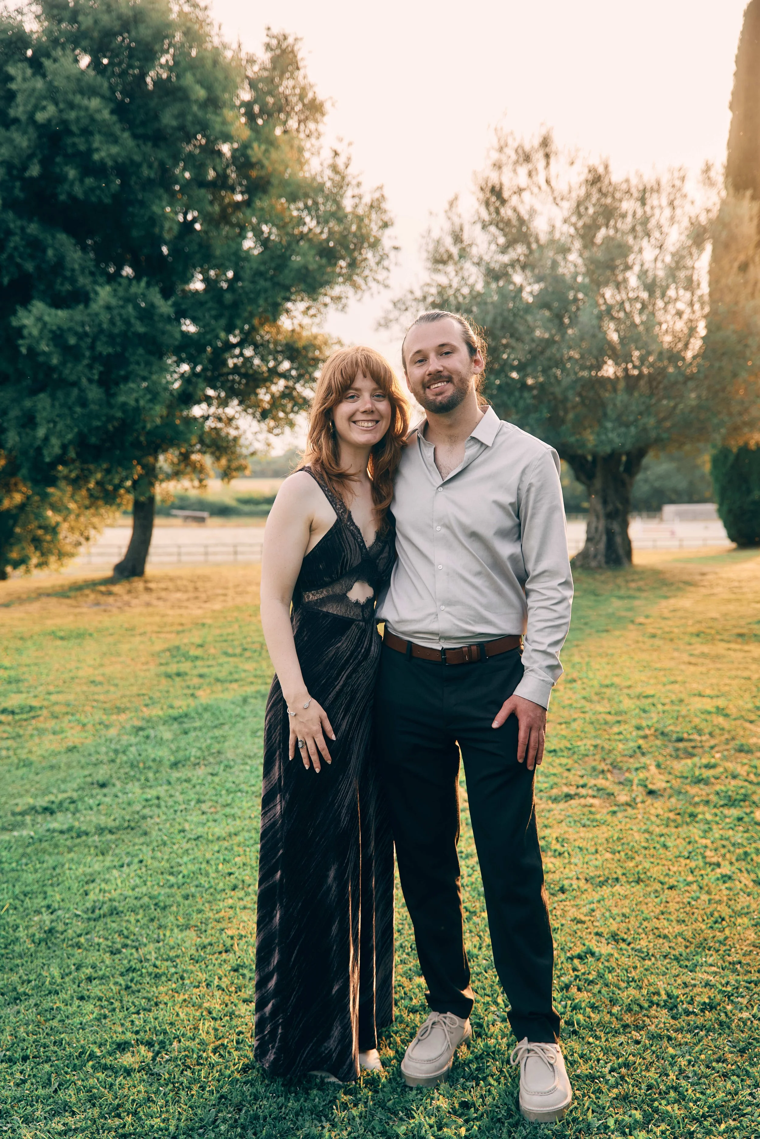 A young couple standing outdoors on a grassy area with trees in the background during sunset. The woman has red hair and is wearing a black dress, and the man has brown hair tied back and is dressed in a light-colored shirt and dark pants.