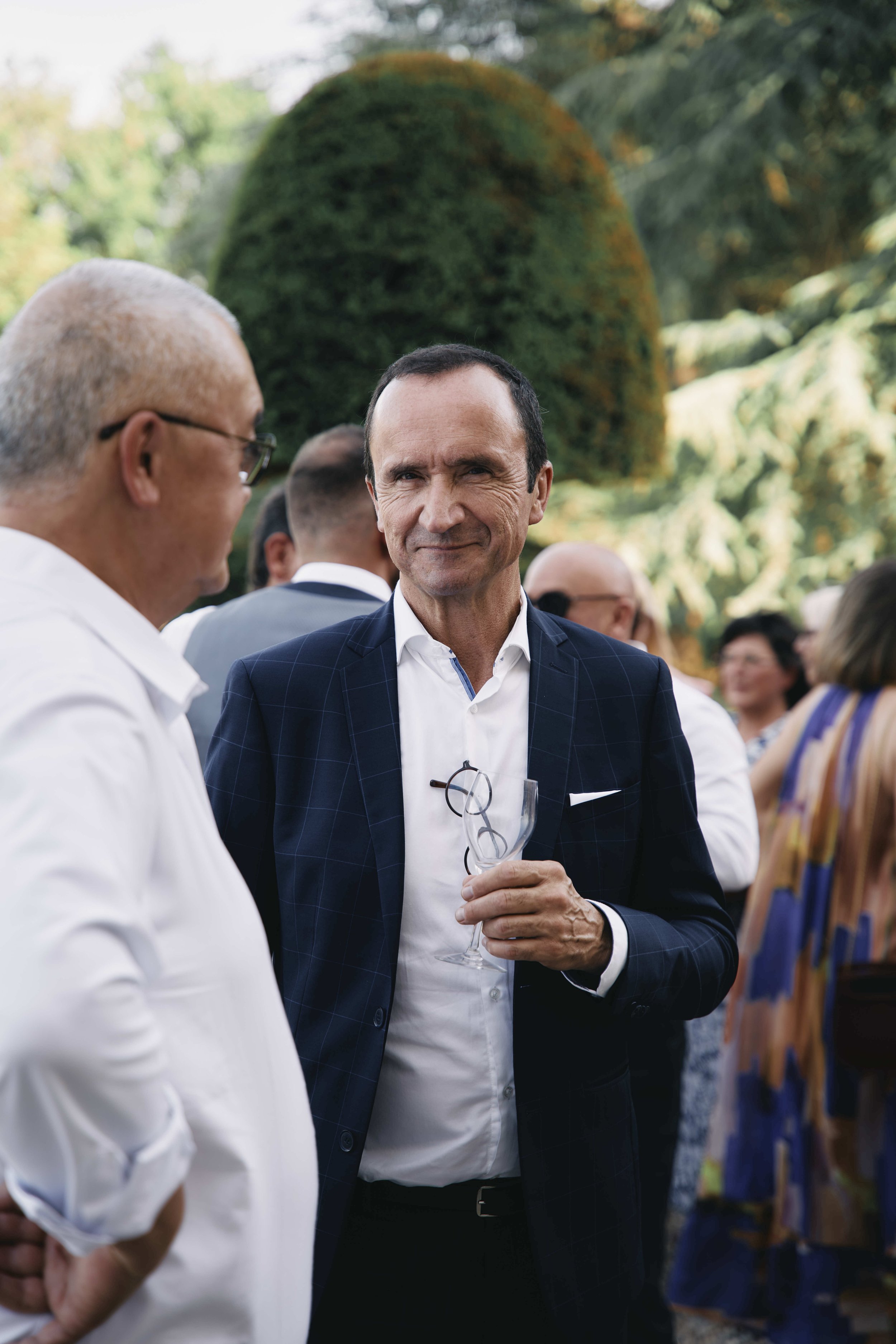 A man in a dark blue blazer holding a glass of champagne, smiling and talking to an older man in glasses. Several people are in the background, outdoors with trees and greenery.