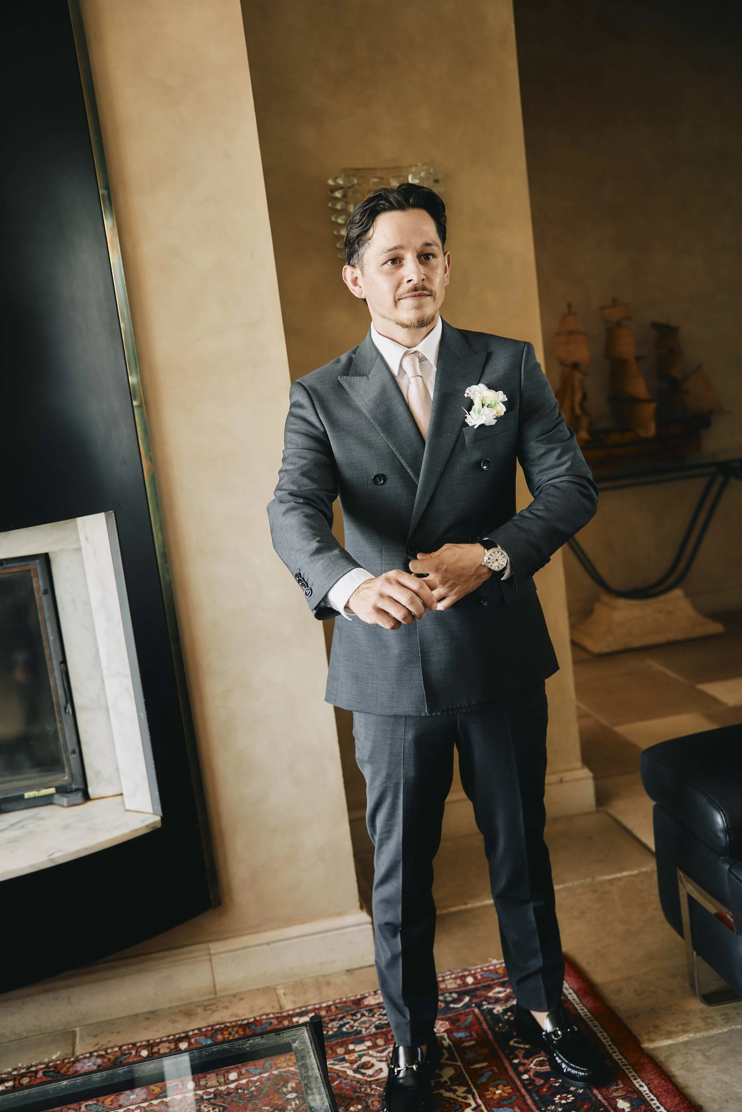 A man dressed in a dark gray suit, white shirt, and cream-colored tie stands indoors against a beige and dark brown wall. He wears a boutonniere on his left lapel and a silver wristwatch on his left wrist. He has short dark hair, a light beard, and l