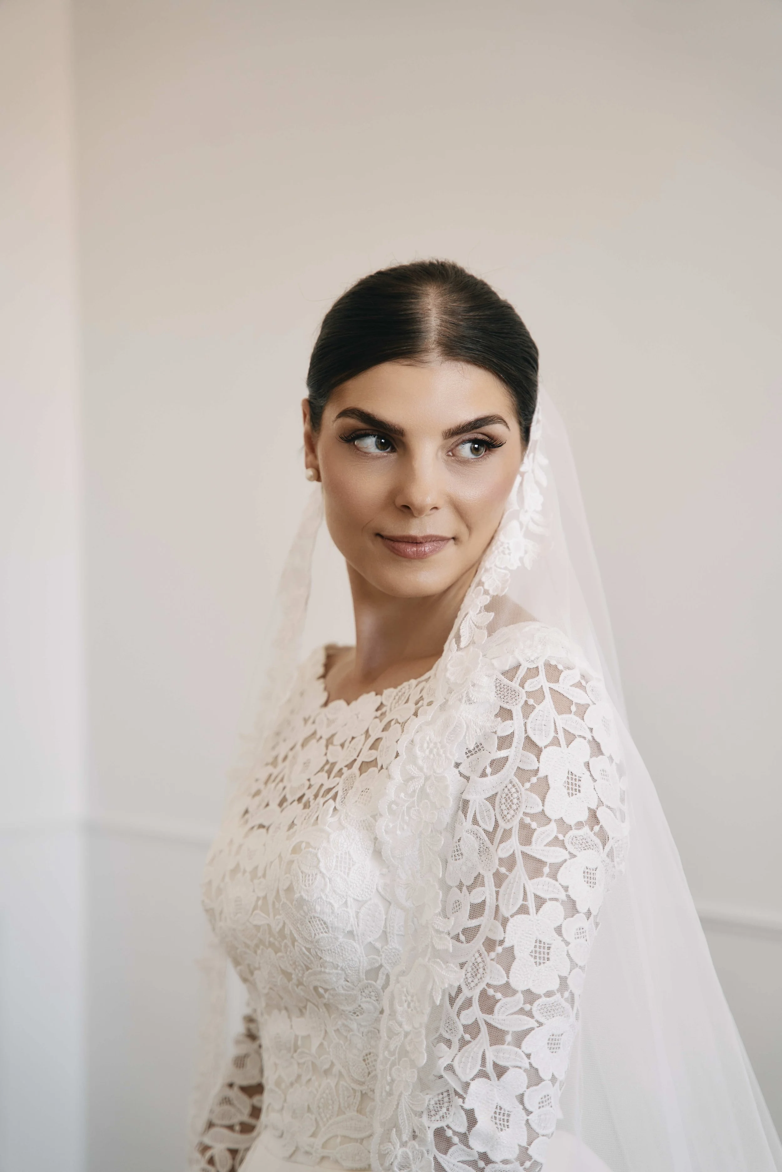 A bride with dark hair styled in an elegant updo, wearing a white lace wedding gown and veil, looking to the side against a plain background.