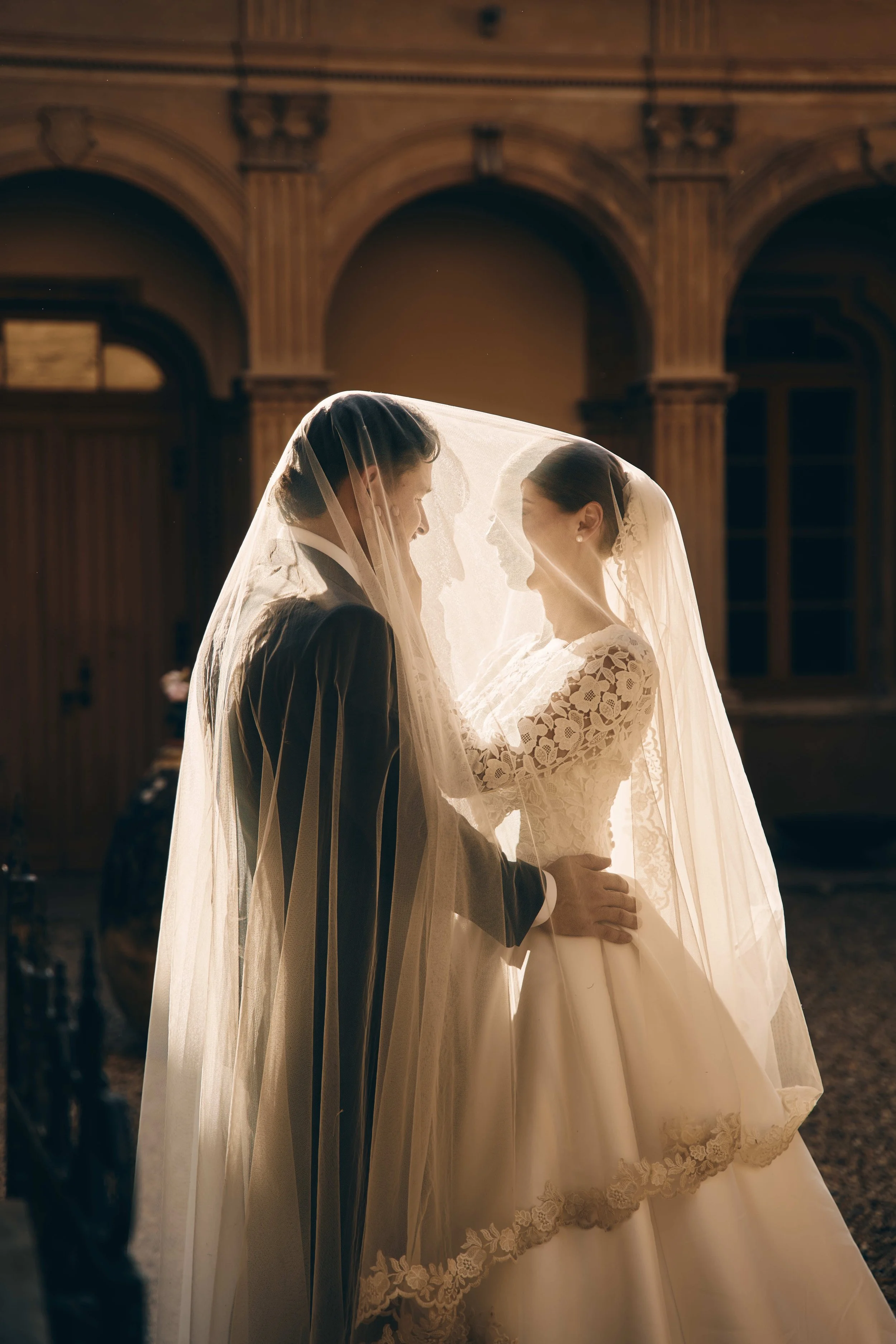 A bride and groom standing close together under a veil in a warmly lit, ornate hall with arched wooden architecture.