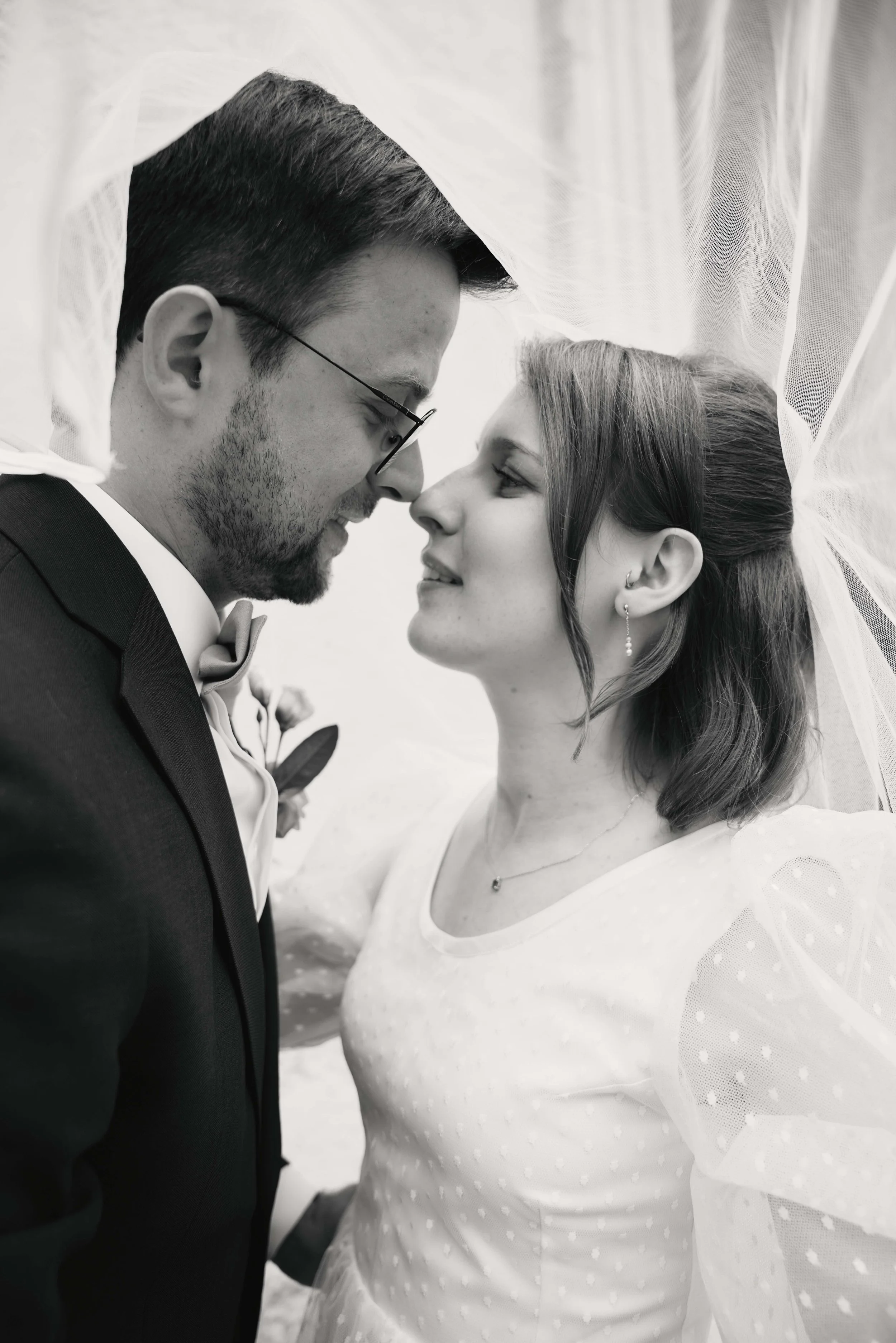 A couple is close together, forehead to forehead, smiling and gazing into each other's eyes, with a sheer fabric curtain draping above them.