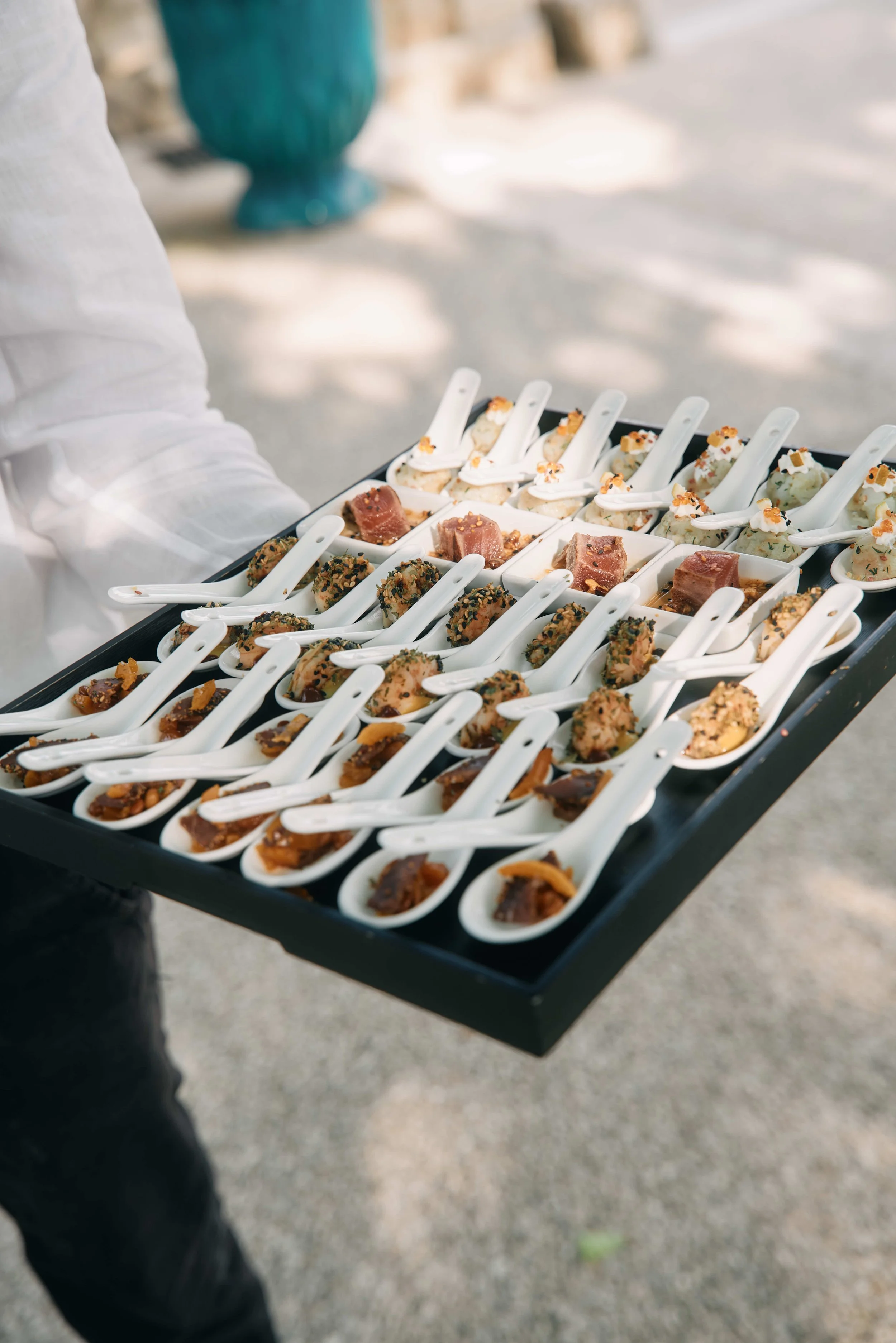 A person in a white long sleeve shirt holding a black tray with small white ceramic spoons filled with appetizers, including some with chopped nuts and others with a creamy topping, outdoors on a sunny day.