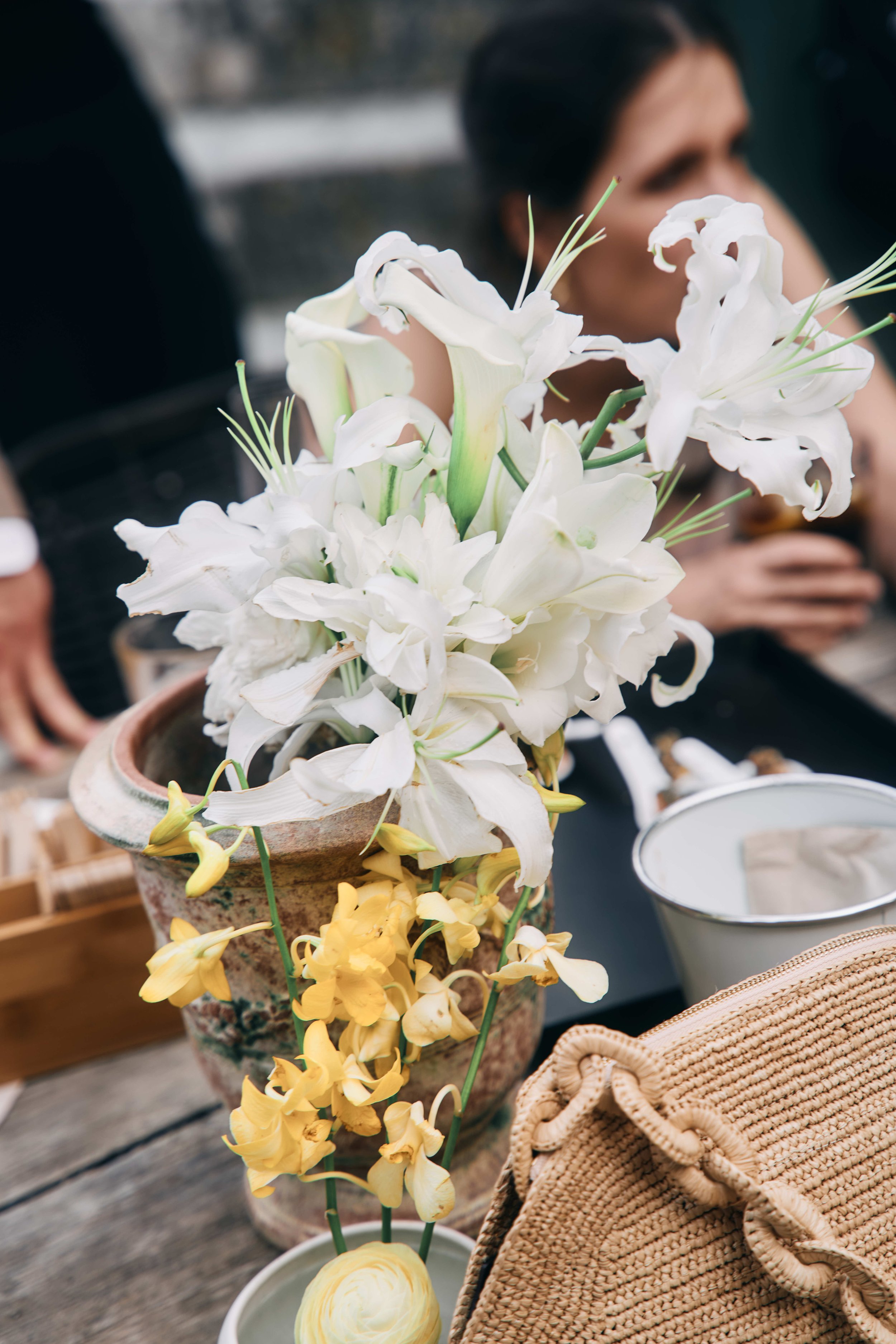 A bouquet of white lilies and yellow orchids in a rustic ceramic pot on a wooden surface with a blurred person in the background.