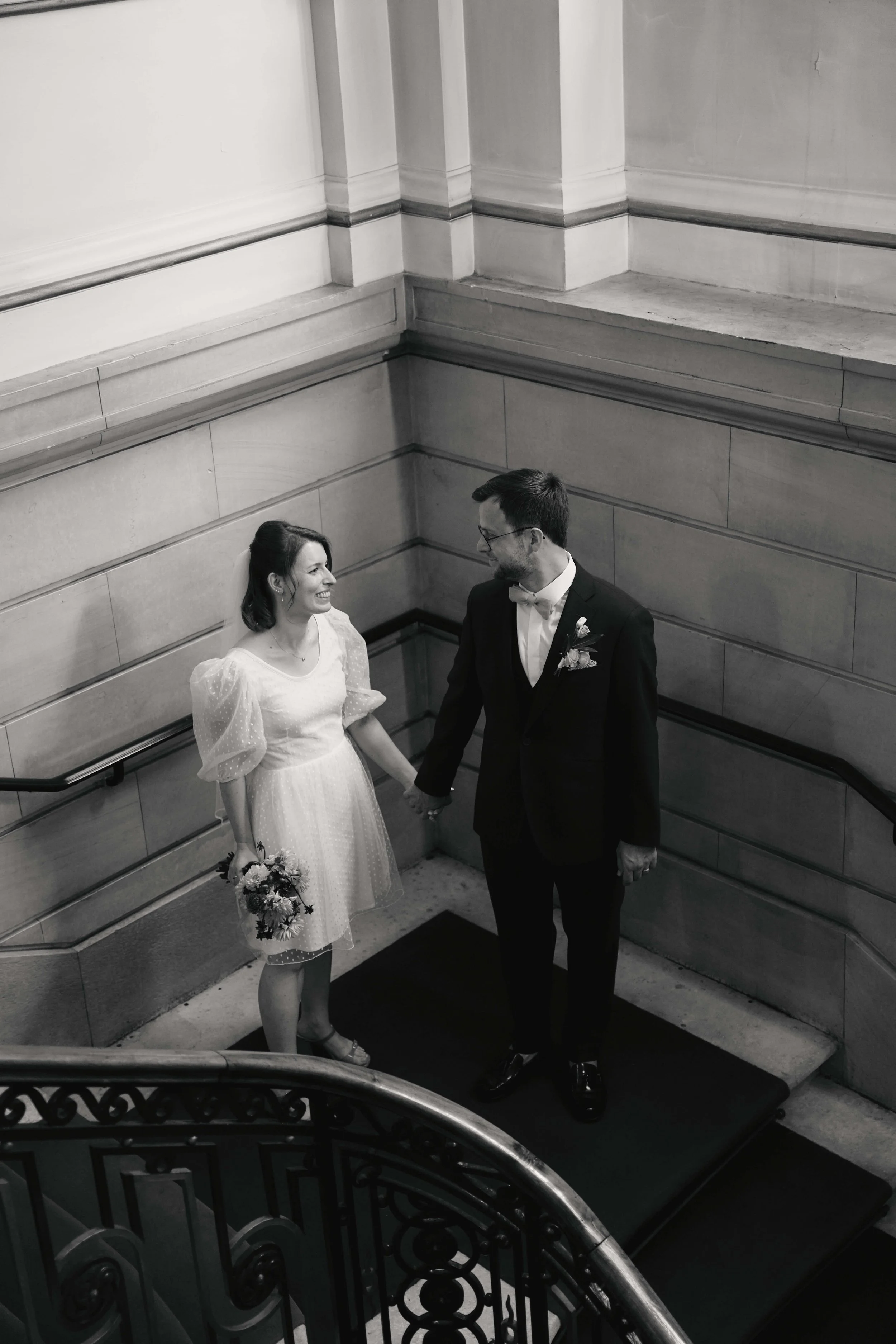 A black and white photo of a couple on a staircase, holding hands and looking at each other. The woman is in a light-colored dress holding a bouquet, and the man is in a tuxedo with a bow tie and boutonniere.