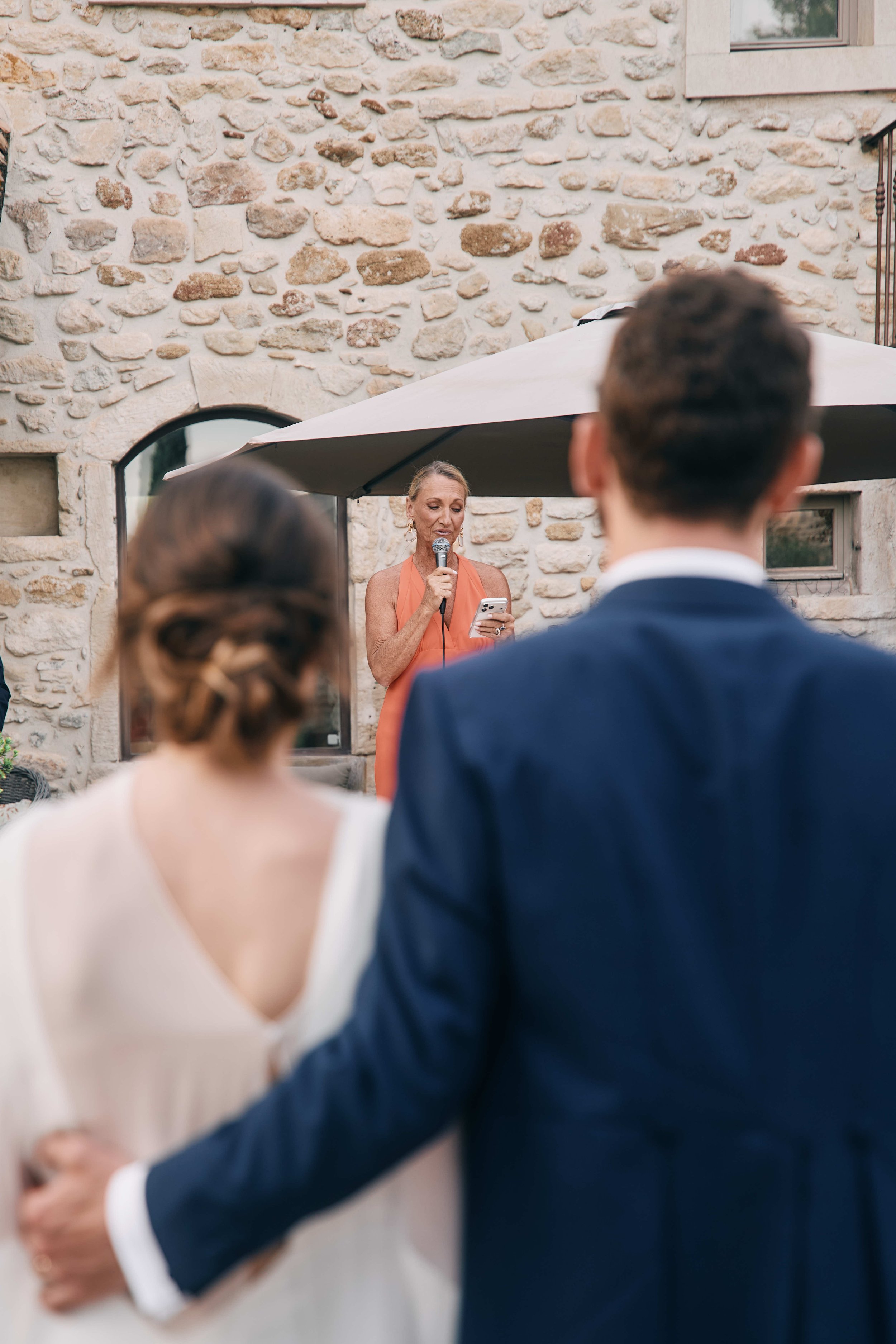 A woman reading from her phone during a wedding ceremony as a man and woman hold each other and listen. The scene is set outdoors against a stone wall with windows, and a large umbrella provides shade.