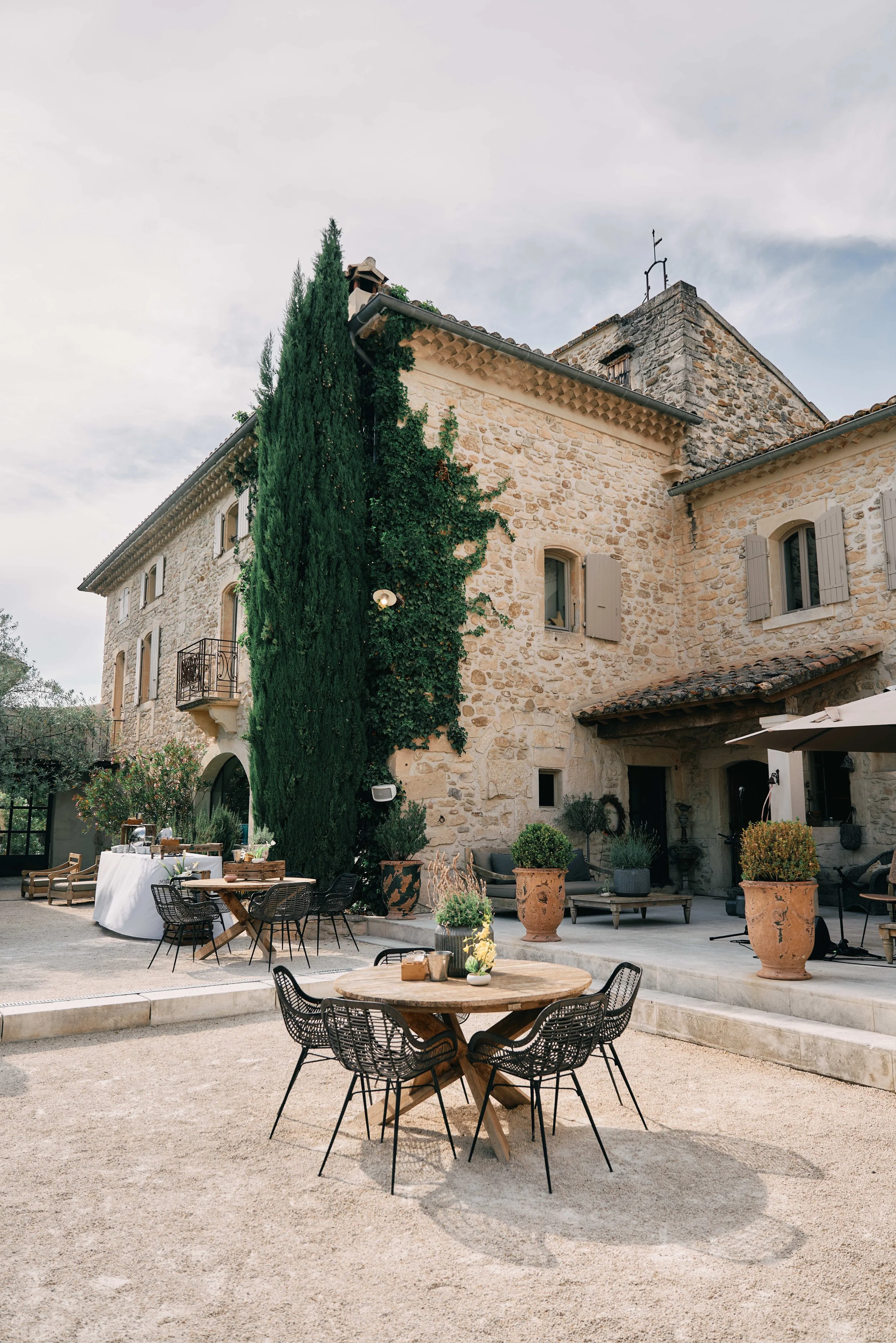 Outdoor patio area with black metal chairs and wooden tables, potted plants, a stone building with windows and closed shutters, and a tall cypress tree under a partly cloudy sky.