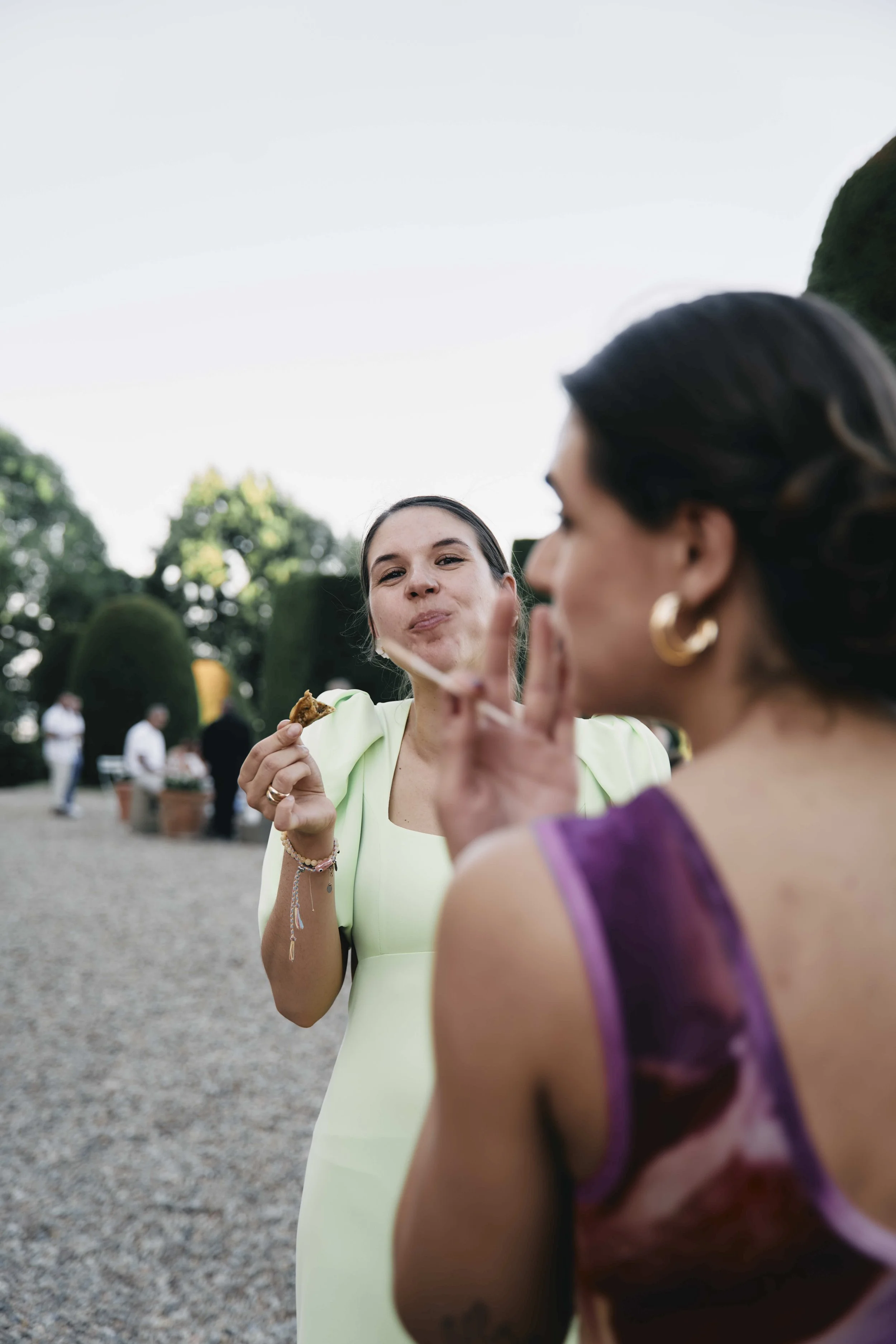 Two women are outdoors at a gathering, one with dark hair, wearing a light yellow dress, holding a slice of pizza and making a finger heart gesture, while the other with dark hair, earrings, and purple clothing, is blurred in the foreground.