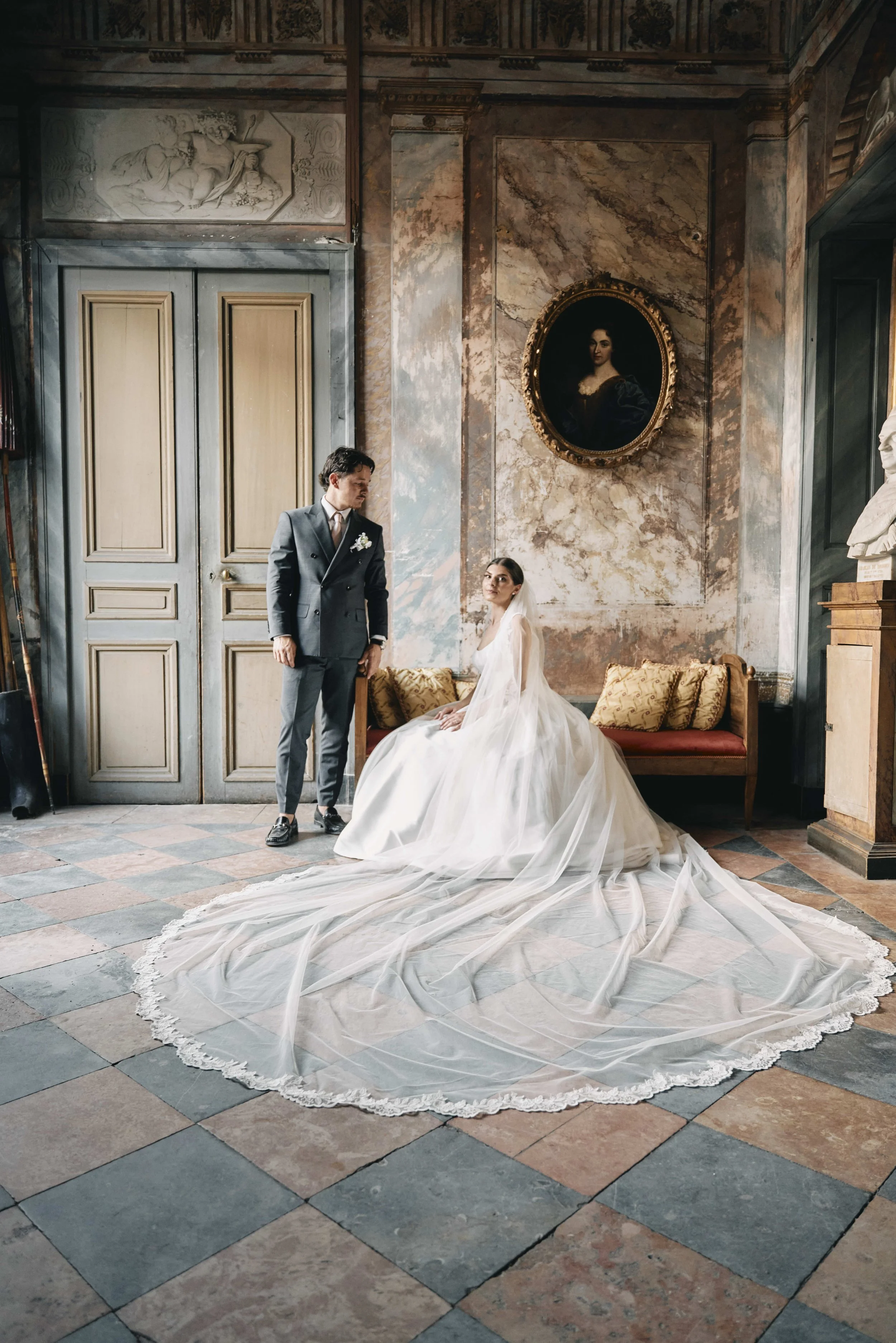 A bride sitting on a vintage sofa with a long bridal train spreading onto the checkered tile floor. A groom stands next to her in a gray suit, inside an ornately decorated, historical interior with large doors, marble walls, and classical art and scu