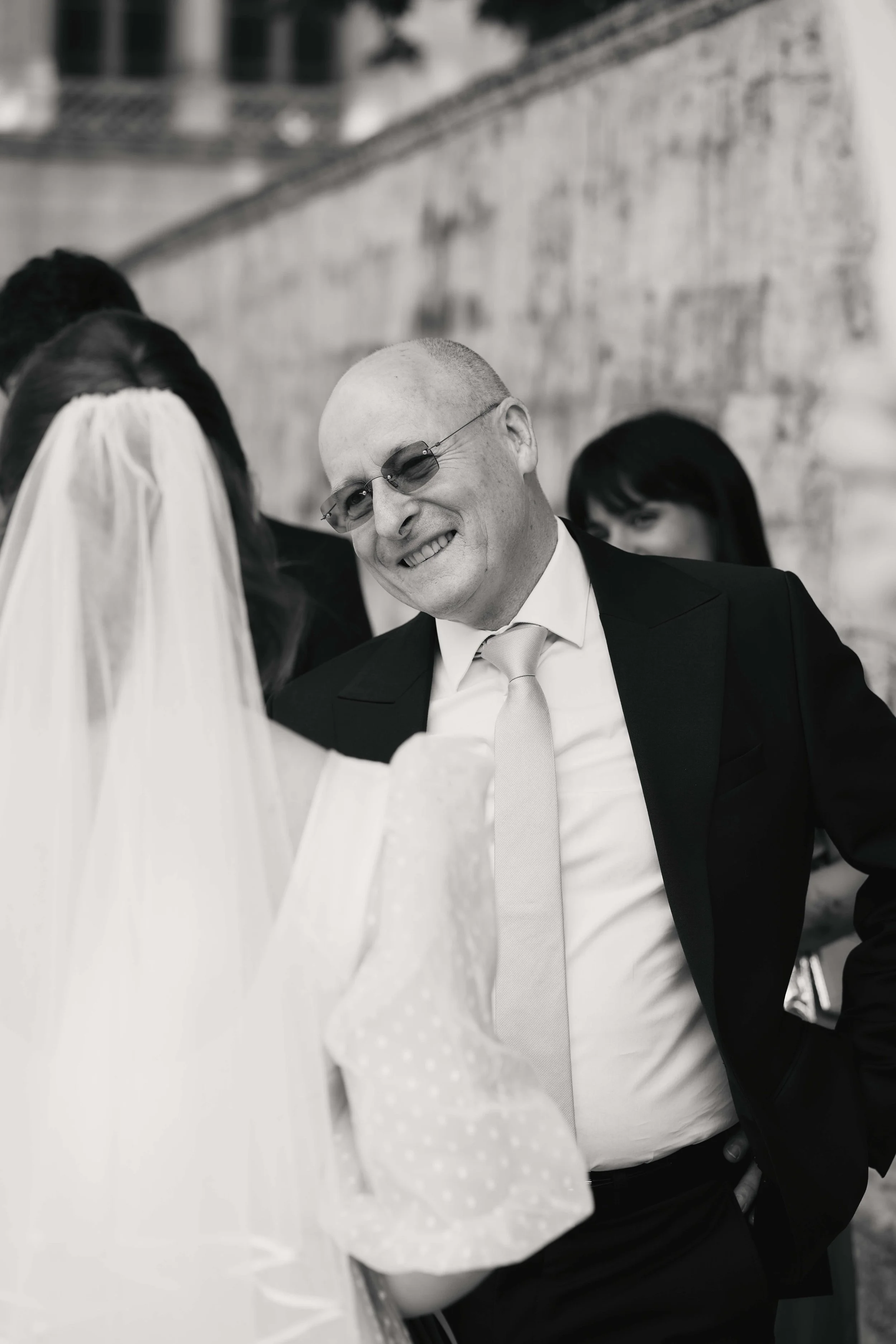 A man with a shaved head and glasses wearing a suit and tie, smiling and looking at a woman in a wedding dress and veil, with other people blurred in the background.
