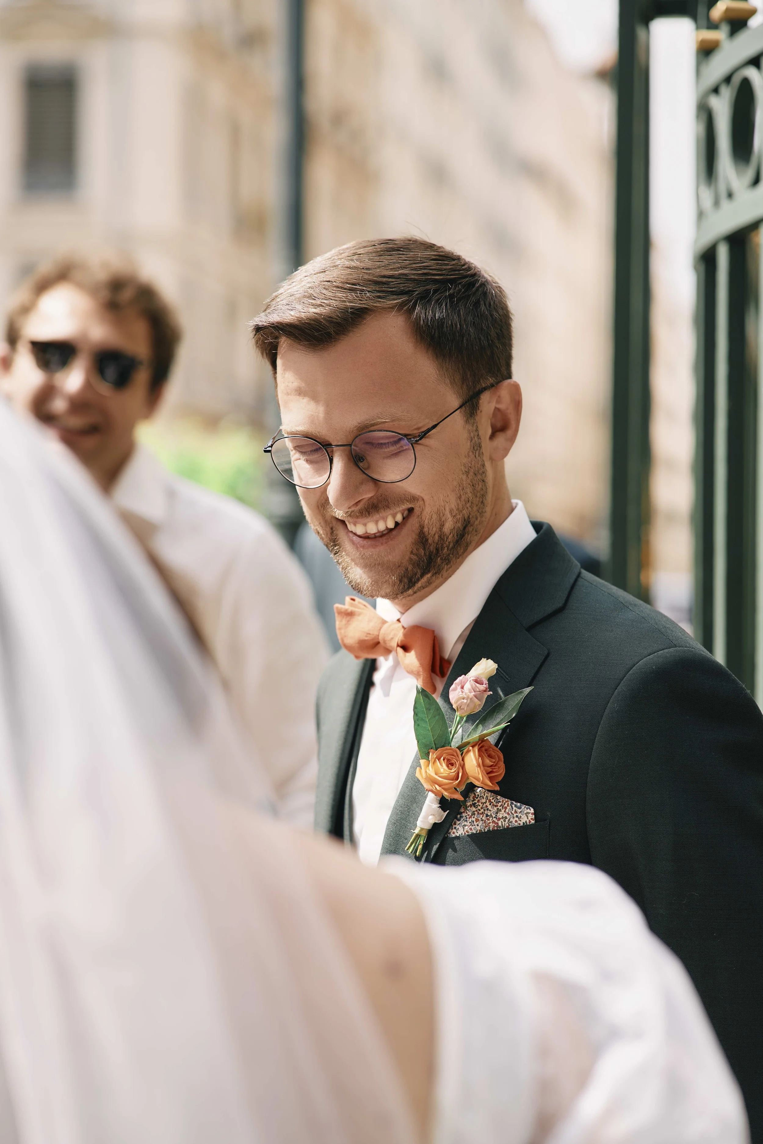 A man wearing glasses, a tuxedo, and a tie with a boutonniere is smiling during an outdoor wedding ceremony. Another man in sunglasses and a white shirt is blurred in the background.